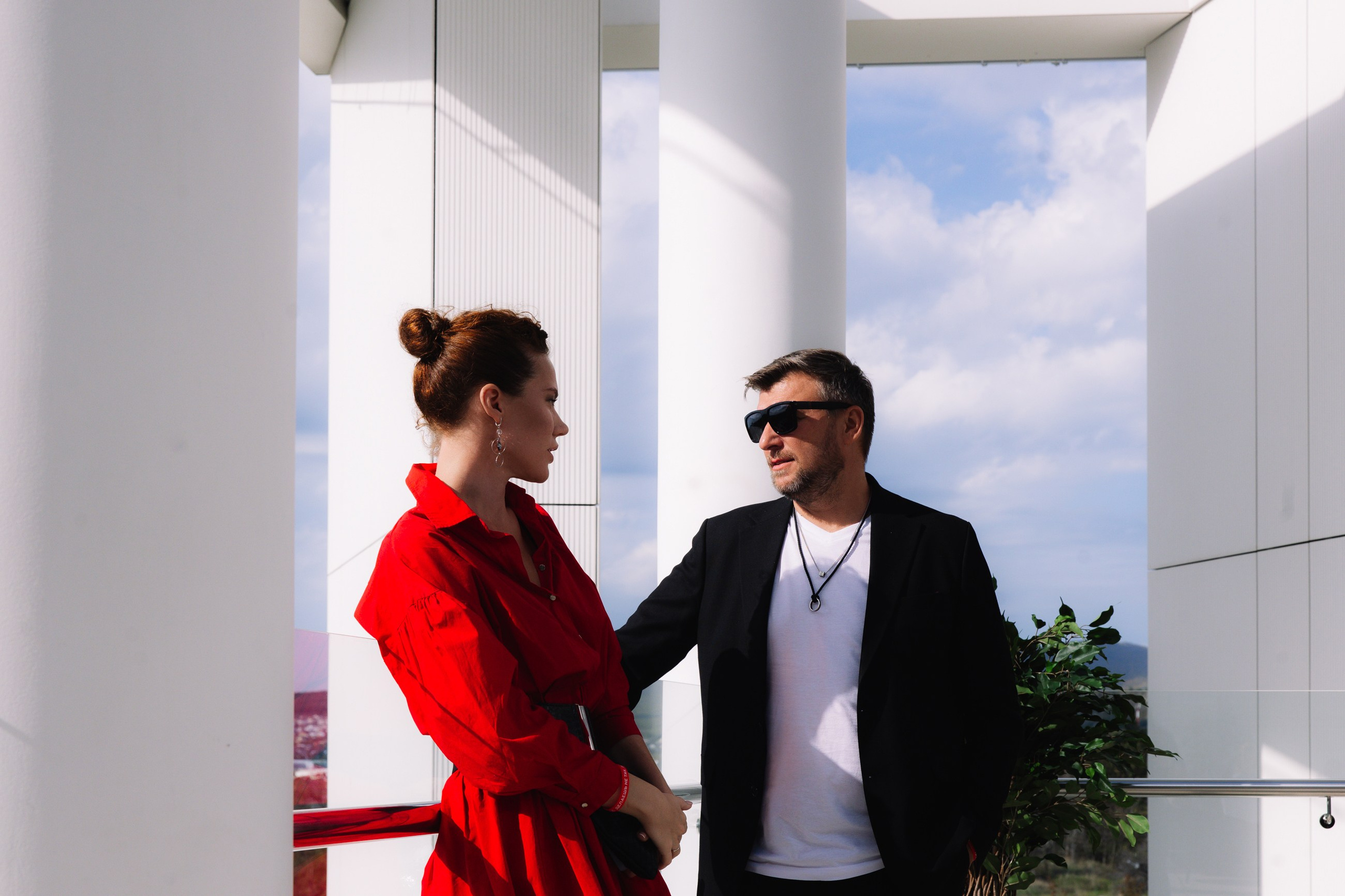 Пара на балконе: Мужчина и женщина в стильных образах (красное пальто и черный костюм) общаются на открытой террасе.