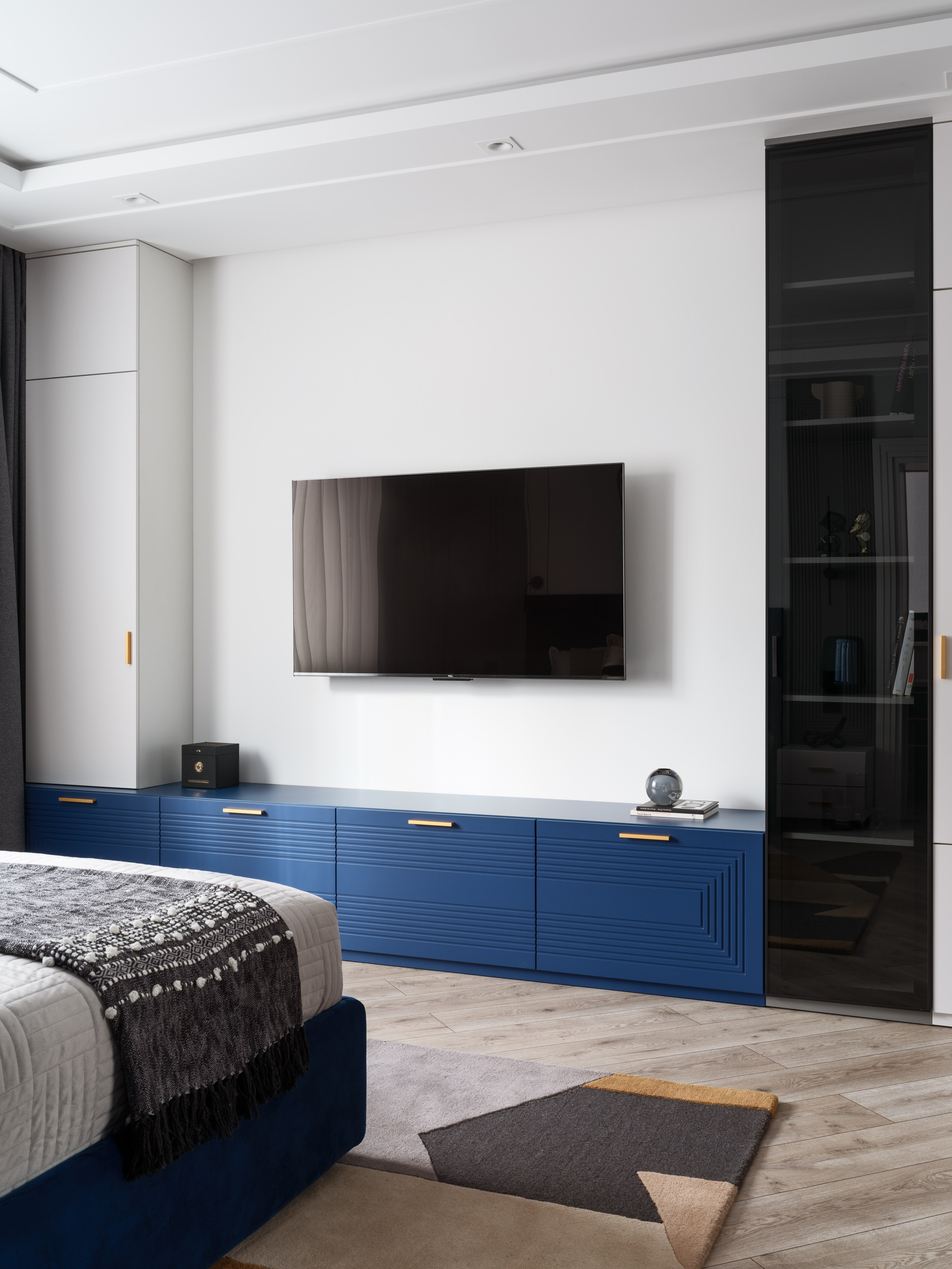 Современный интерьер спальни с минималистичной композицией: белая стена с телевизором, стильная тумба с глубокими синими фасадами и золотыми ручками. Проект создан дизайнером Евгенией Ермошиной, профессиональное фото Сергея Денисенко.