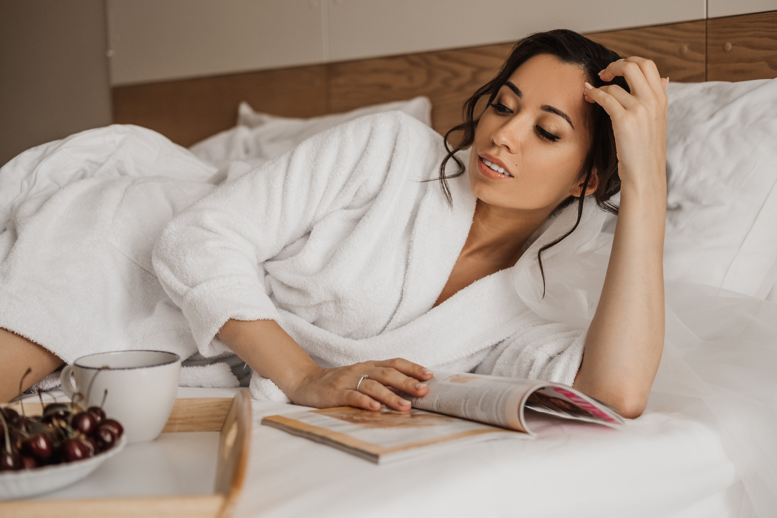 Девушка в белом халате читает модный журнал на кровати.