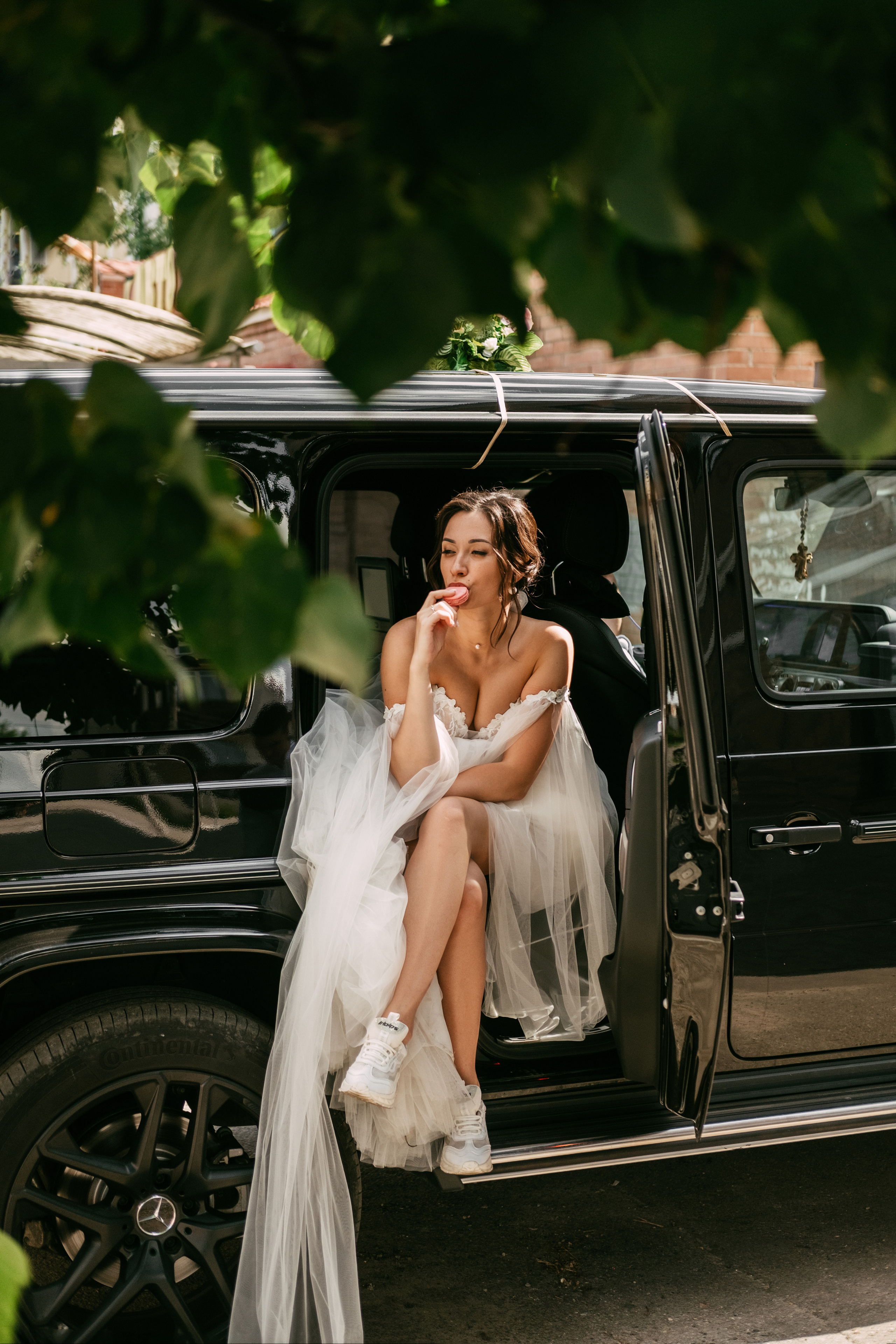 Девушка в свадебном платье и белых кроссовках сидит в машине и перекусывает.