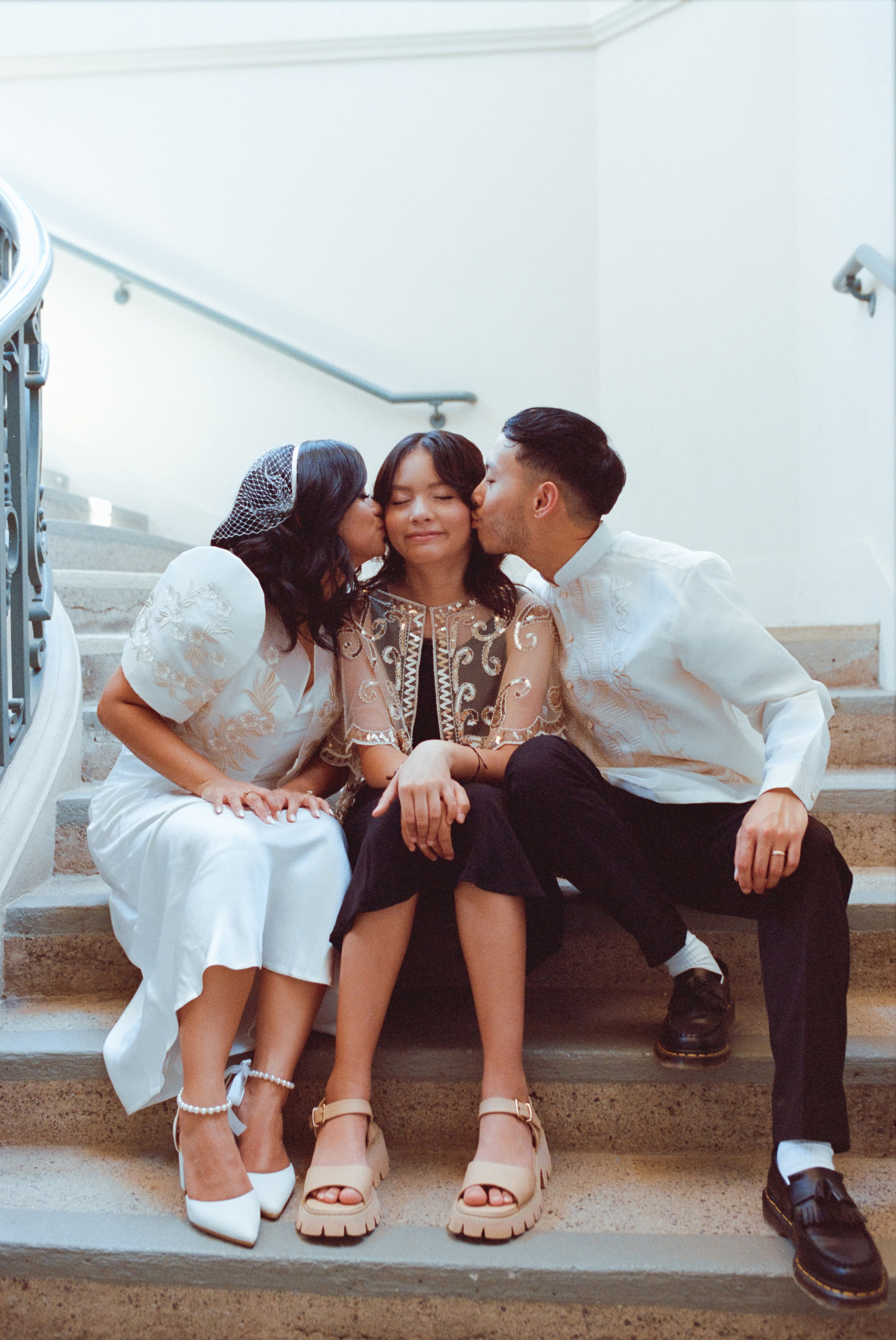 a bride and groom kissing their daughter and sitting on the stairs in the city hall