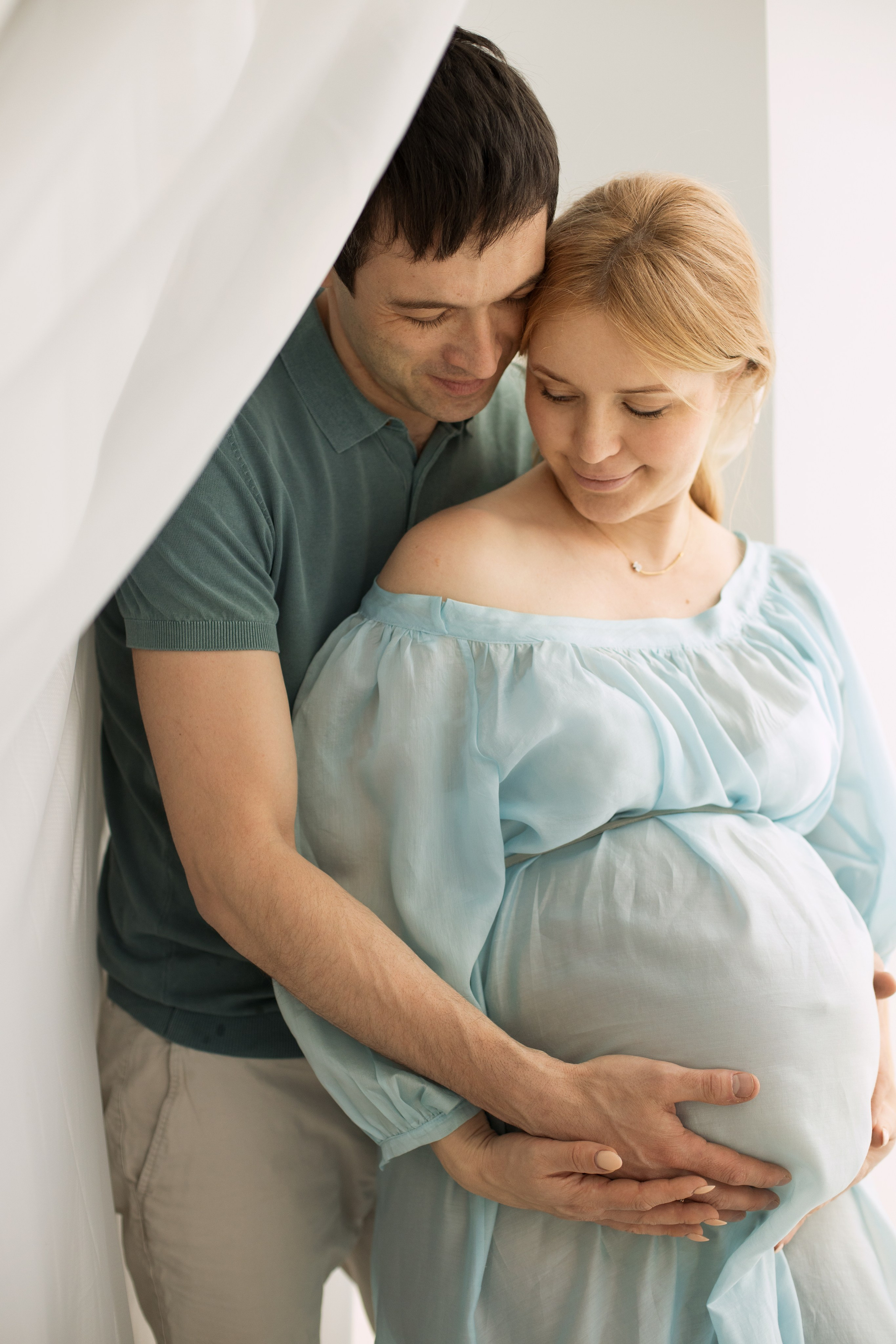 Муж обнимает беременную жену в голубом платье, стоя у окна.
