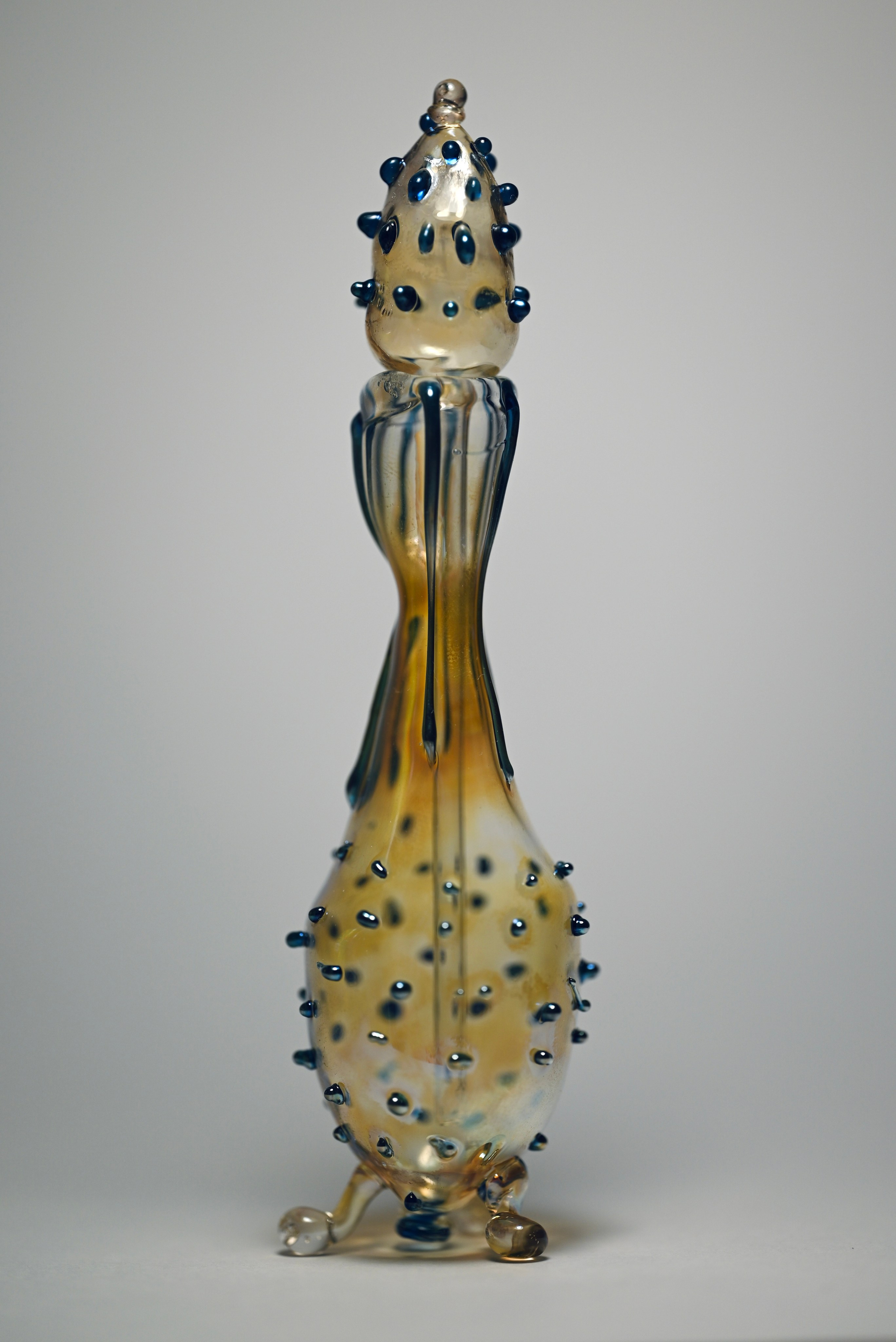 alt="Стеклянная скульптура в виде вытянутой бутылки с золотистым оттенком и округлыми выступами по всей поверхности."
