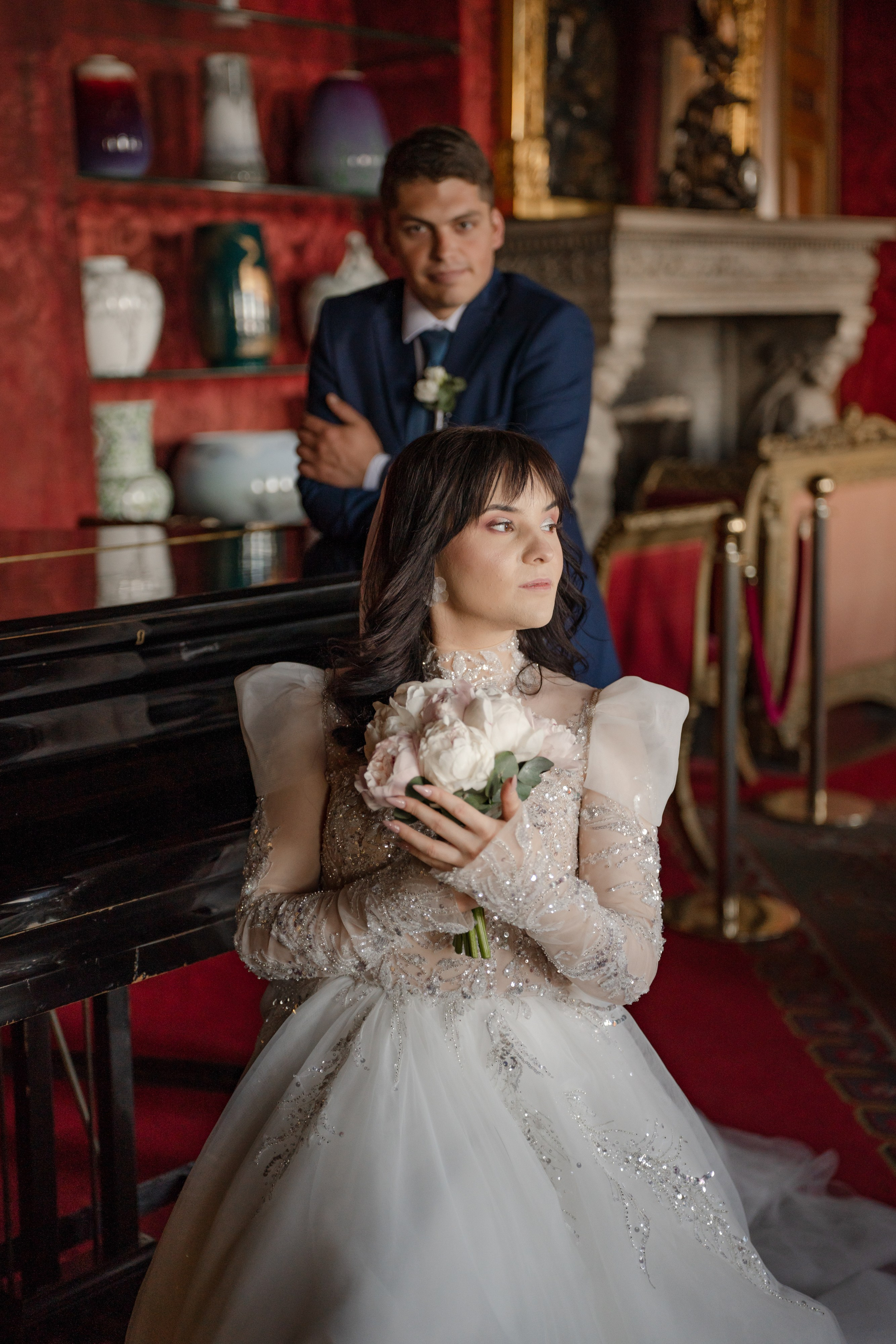 Свадебная фотосессия в Доме ученых, Дворце князя Владимира в Санкт-Петербурге. Невеста сидит на стуле, жених стоит рядом в красной гостиной.