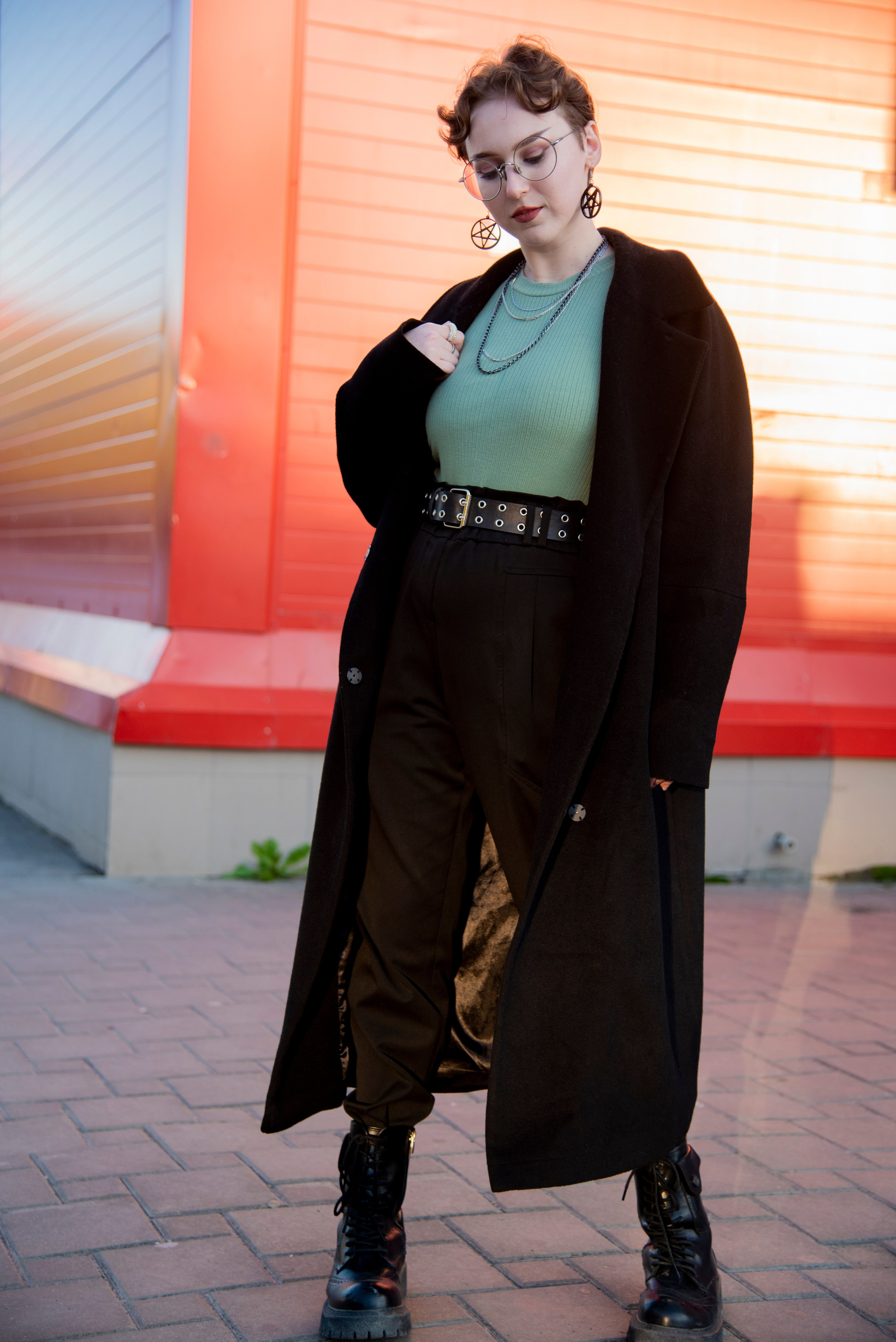 портет девушки. чёрное пальто. красивые позы. серьки в виде пентограммы. короткая стрижка. круглые очки. красный фон