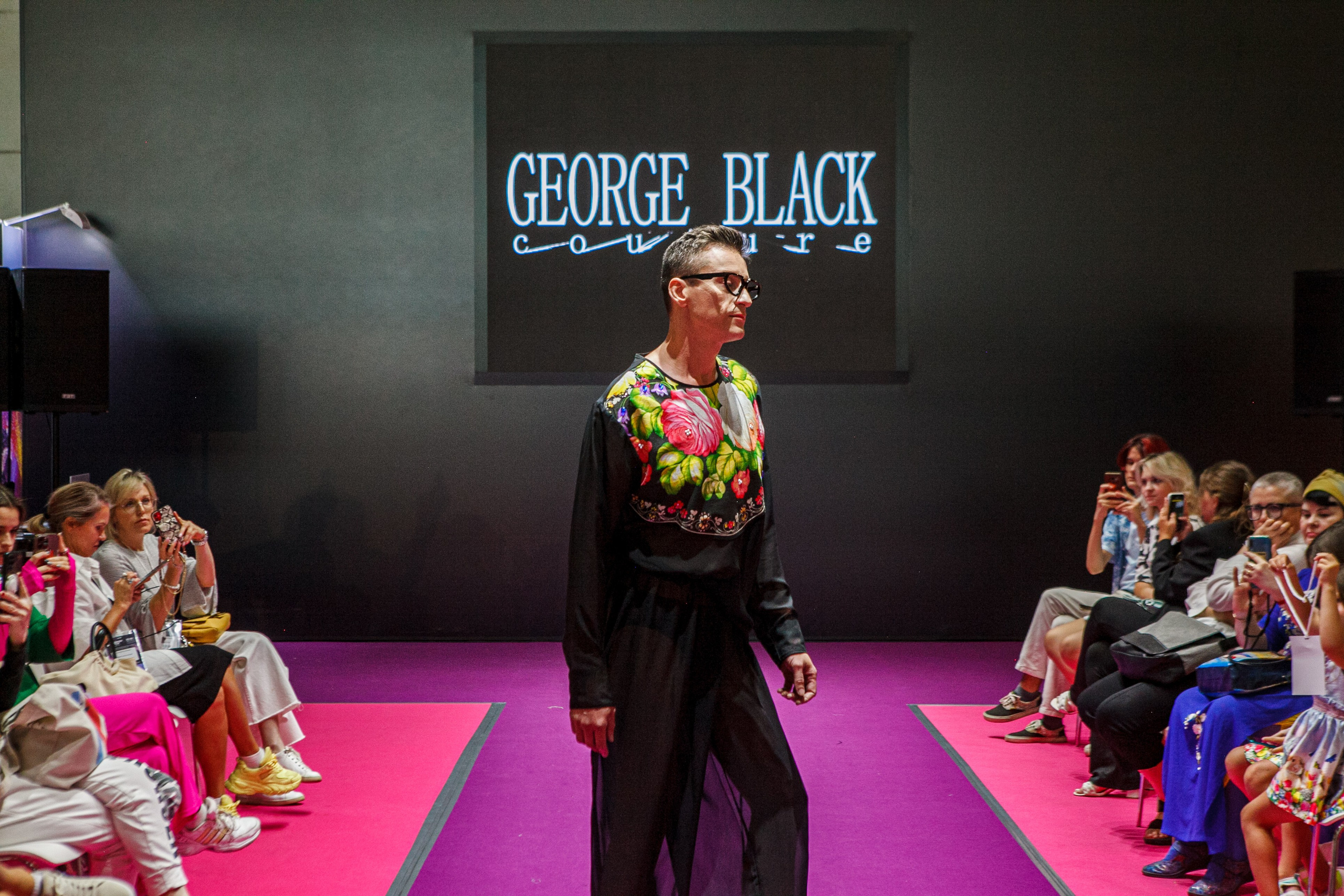  "Модели в ярких нарядах проходят по подиуму во время показа мод, освещенные яркими софитами и окруженные восторженными зрителями."