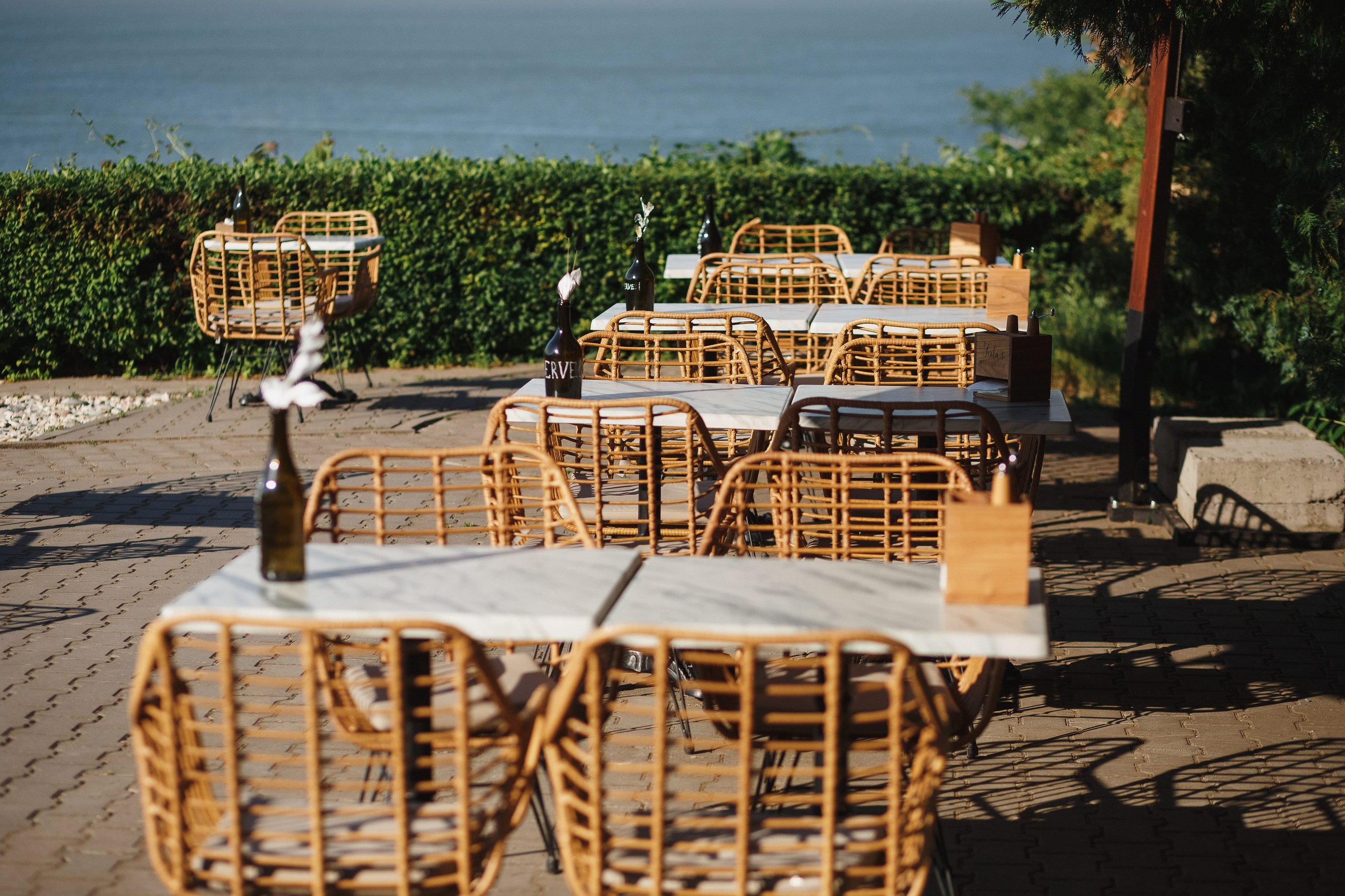 Открытая терраса ресторана Feola's Kitchen & Wine в Таганроге с плетеными креслами и столиками, на фоне зелёных кустарников и Таганрогского залива.