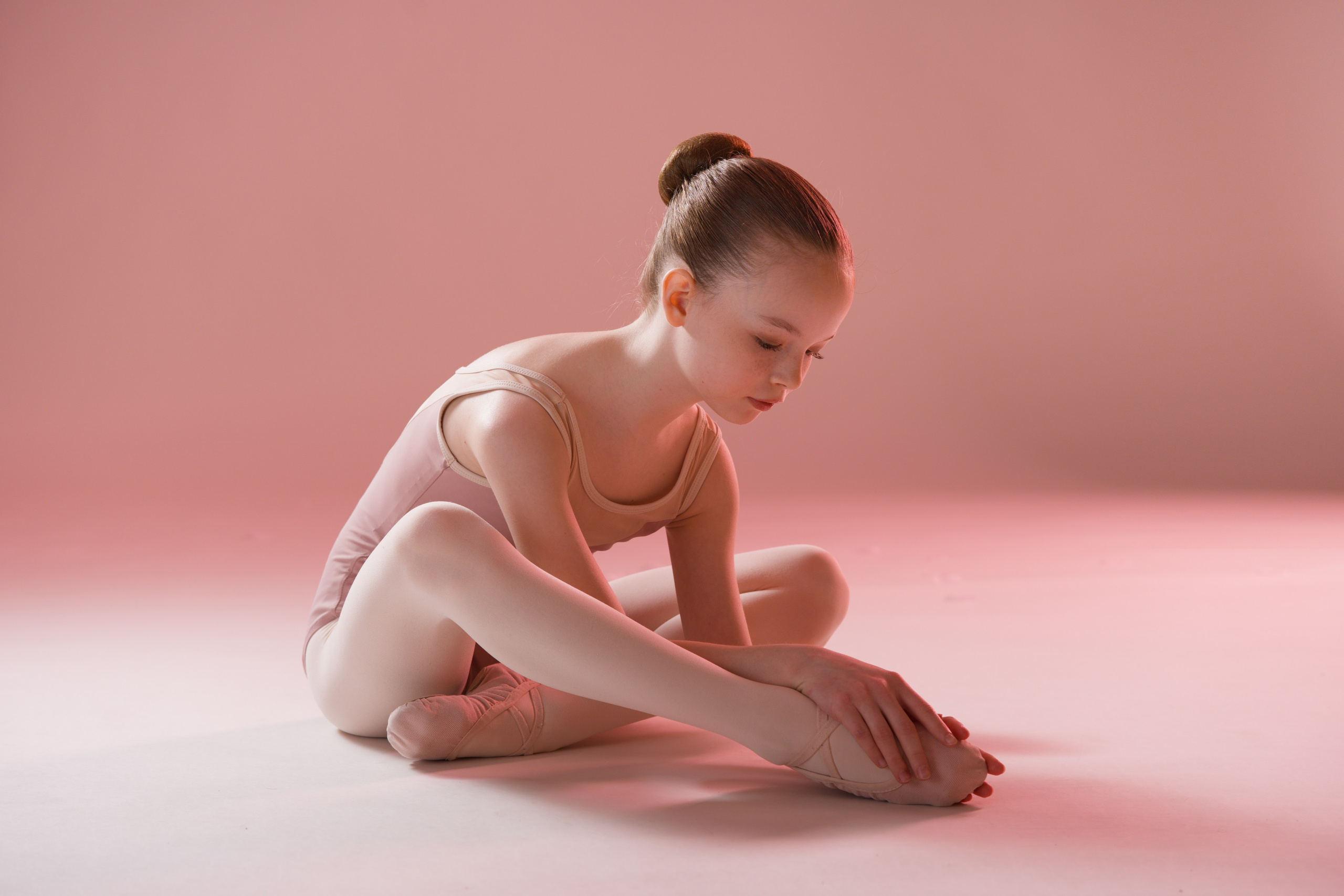 Балетная поза сидя с наклоном к стопе, мягкое студийное освещение в пастельных тонах, художественная серия «Воздушность» в Тюмени