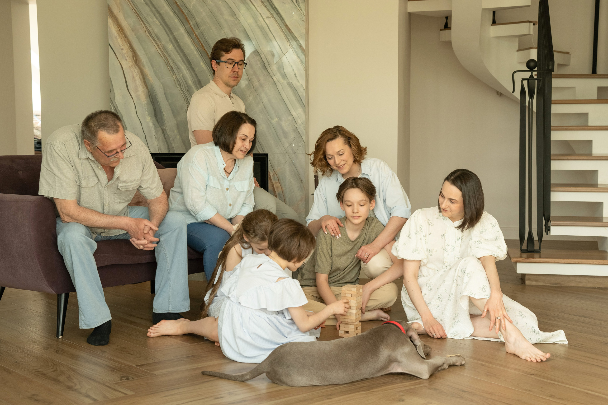 Большая семья играет в настольную игру. Все одеты в светлые одежды. Им весело и они смеются. Семья состоит из бабушки, дедушки, тети, мамы, папы, трех детей.