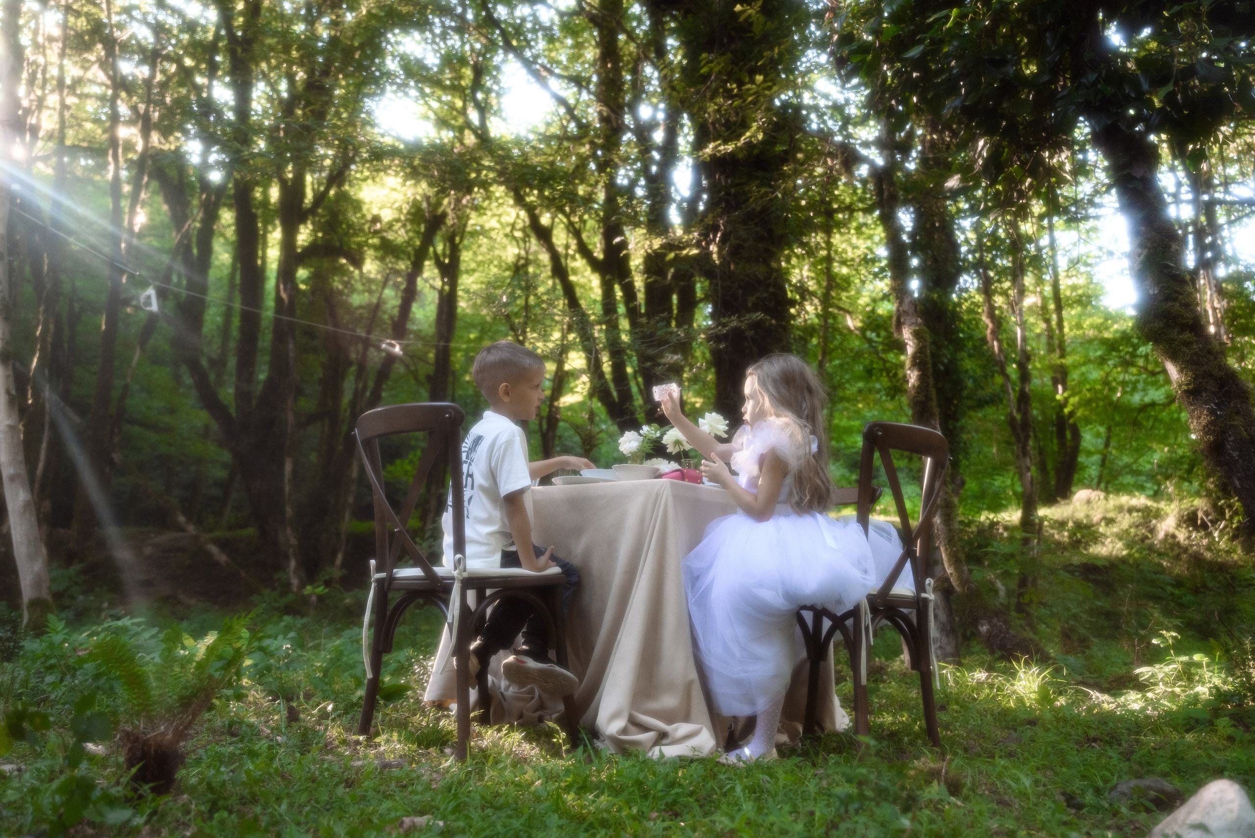 Лес слева пробивается луч солнца который попадает в объектив и создает красивый блик в центре кадра стол за котором сидят двое детей девочка и мальчик они что-то рассказывают друг другу 