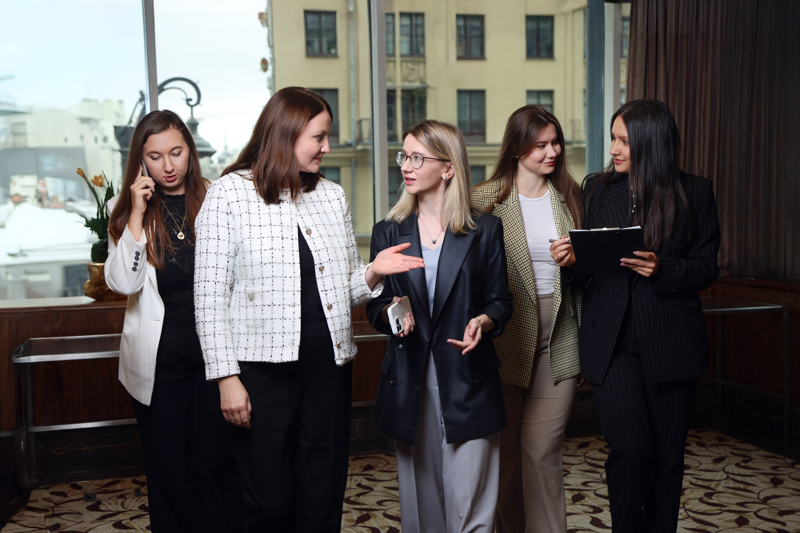 Группа из пяти сотрудниц отеля (из разных отделов) идет по коридору, обсуждая рабочие задачи. Командное взаимодействие.