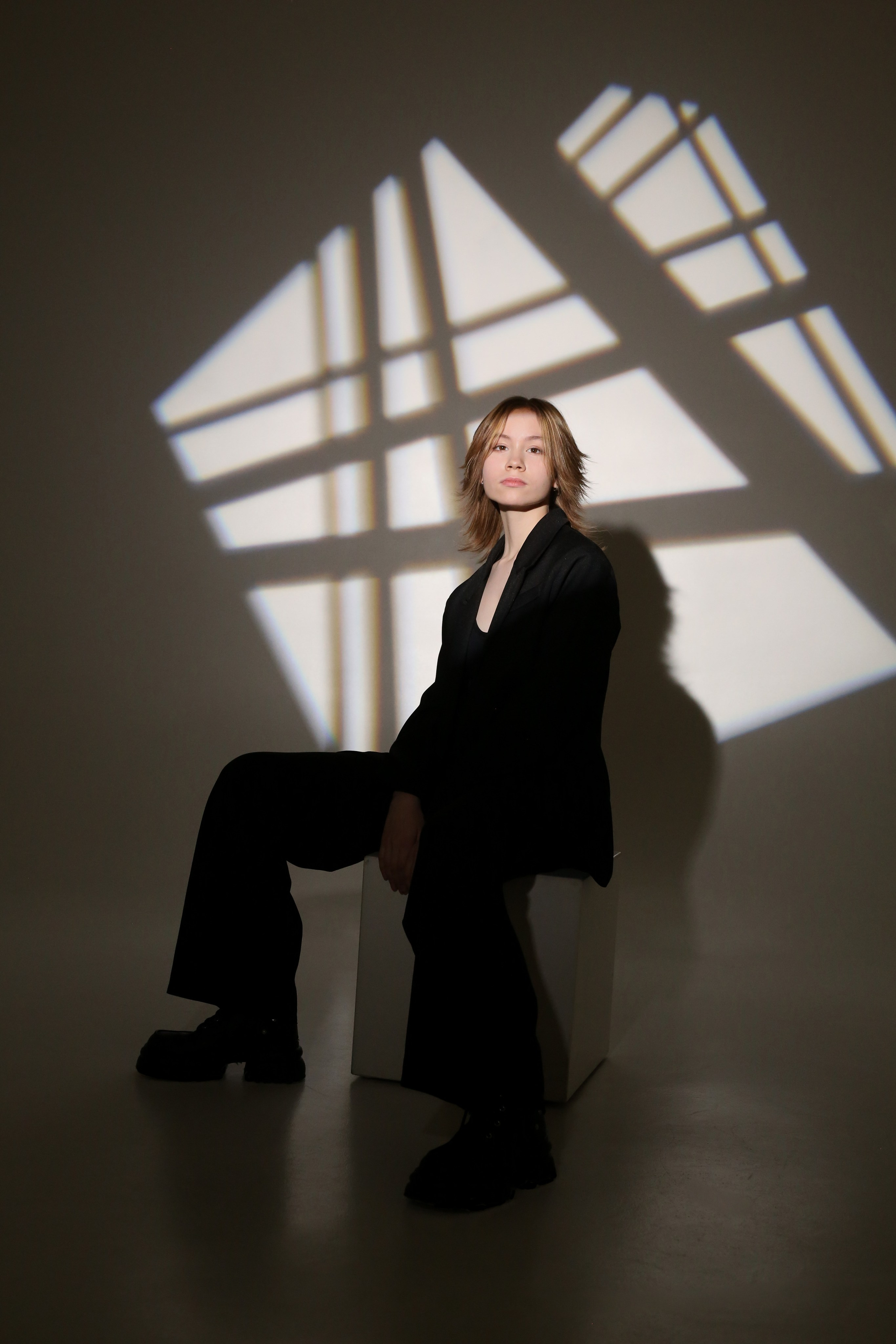 Портрет из фотосессии «Геометрия»: девушка в черном костюме сидит на кубе в лучах света