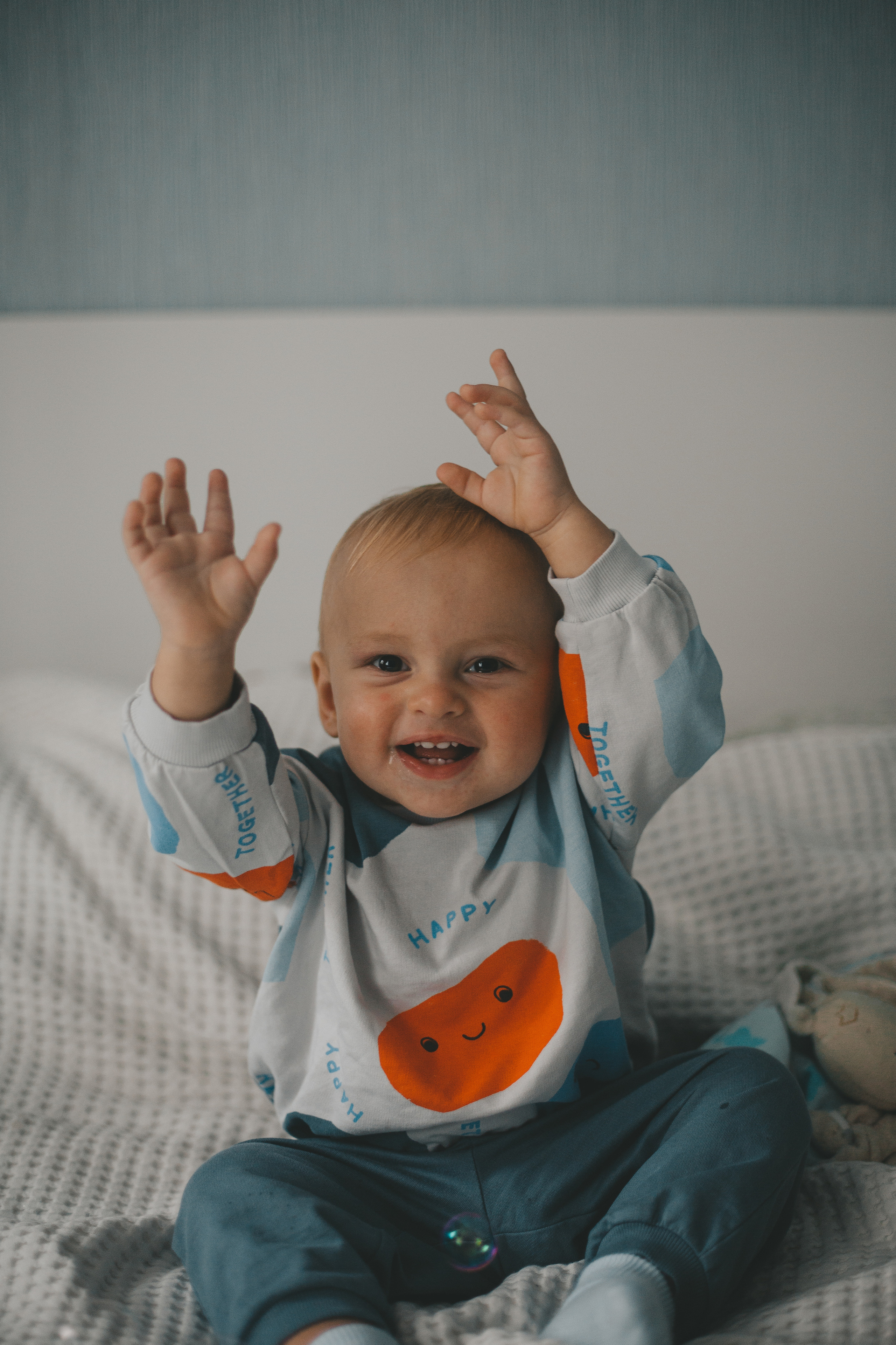 малыш улыбается, годовалый ребенок сидит на кровати и смеется