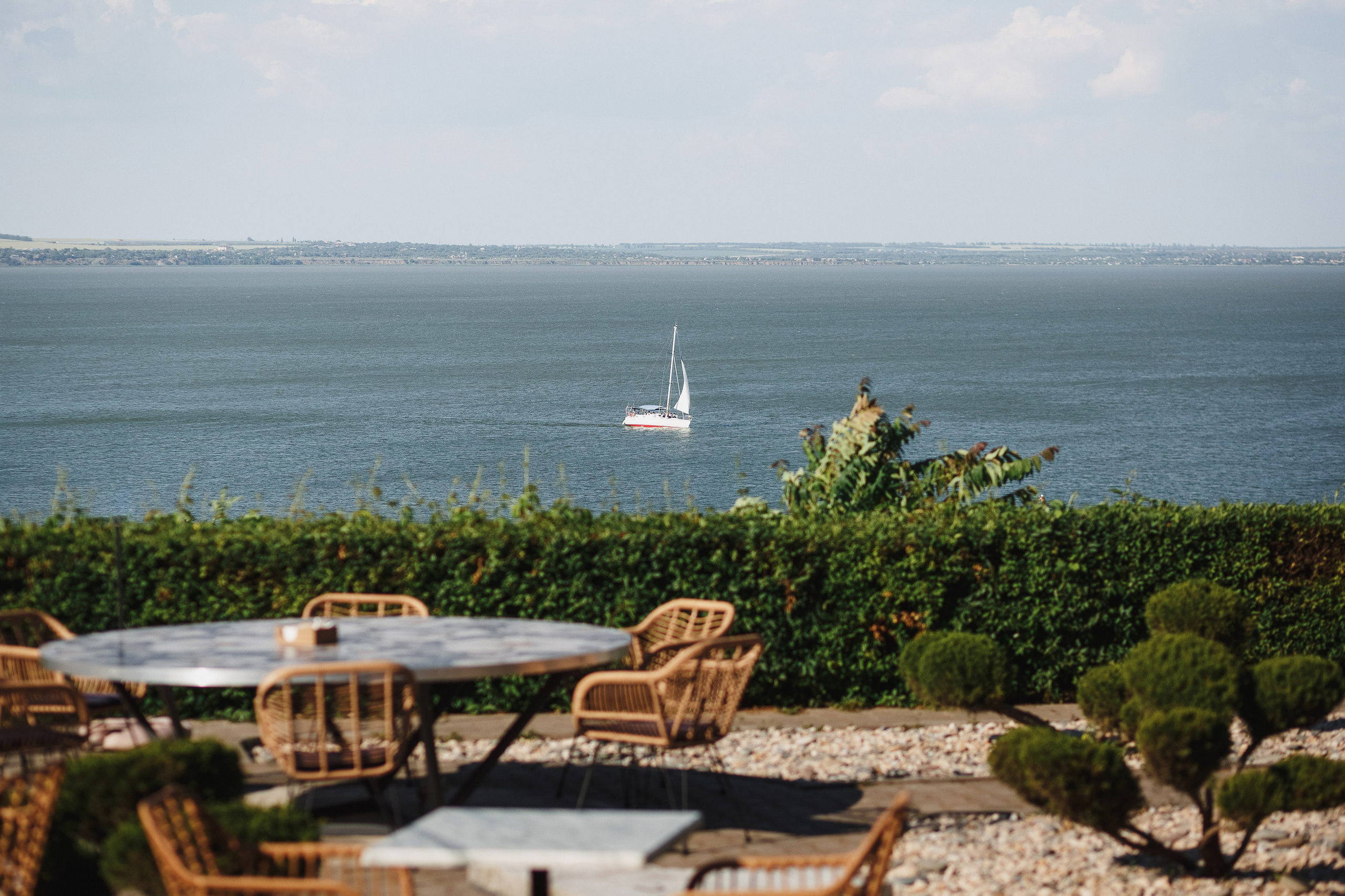 Вид на Таганрогский залив с террасы ресторана Feola's Kitchen & Wine в Таганроге. На переднем плане столы и стулья на открытом воздухе, а на воде видна белая яхта с парусами.