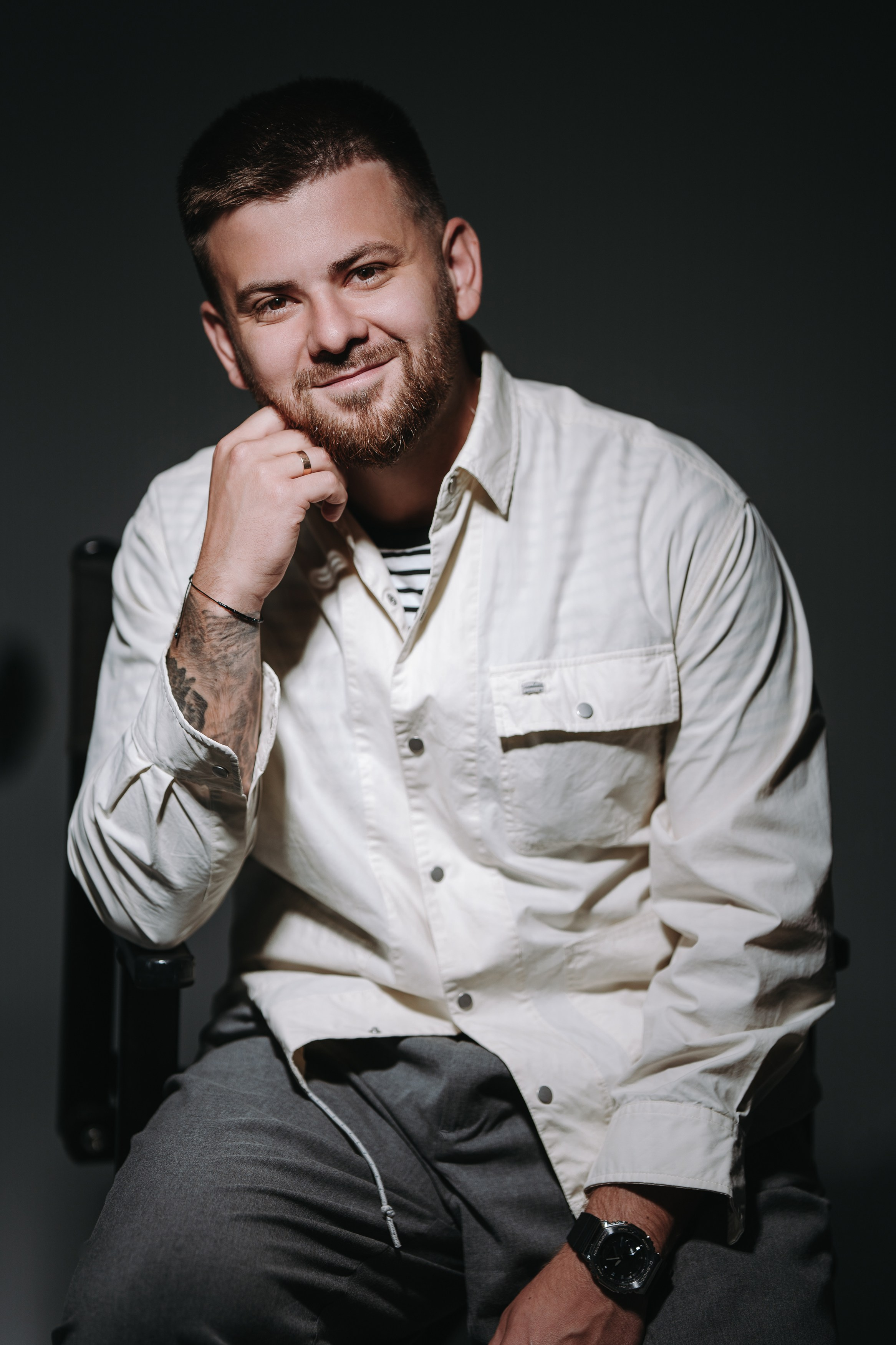 Бородатый мужчина в светлой рубашке и тельняшке сидит на стуле, опираясь на руку и улыбаясь. Тёмный фон и мягкий свет создают стильный портрет