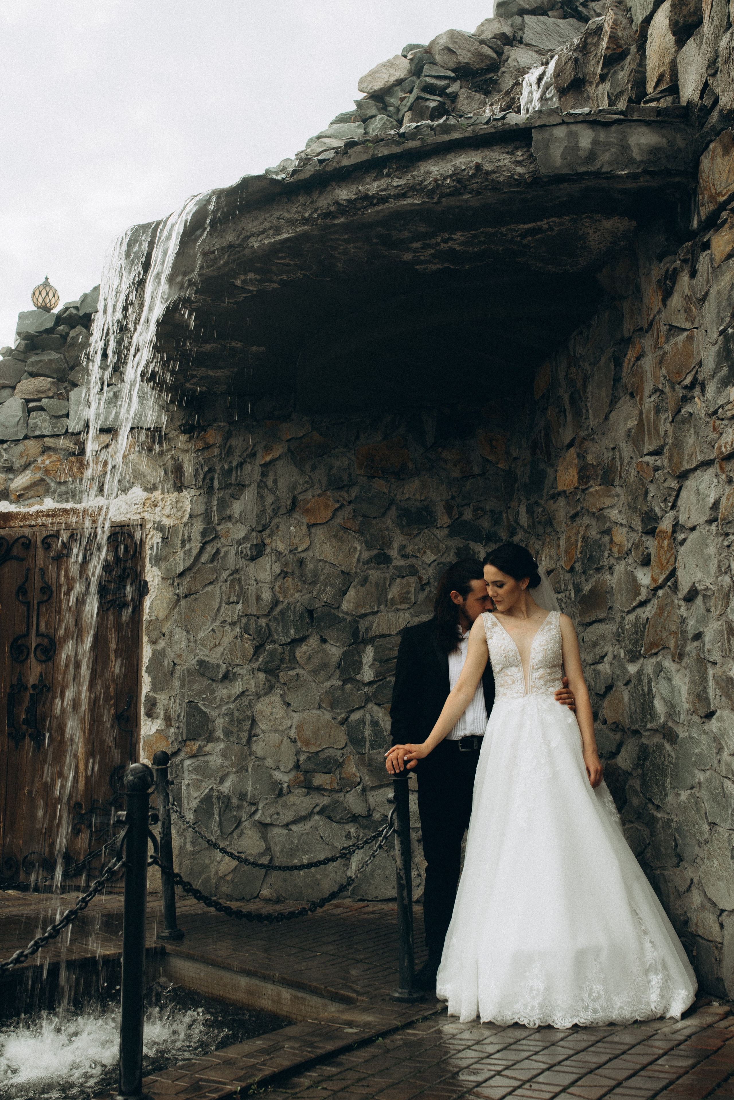 Жених Никита держит невесту Линду за руку, обнимая за талию, у каменного водопада. Свадебная фотография в загородном комплексе.