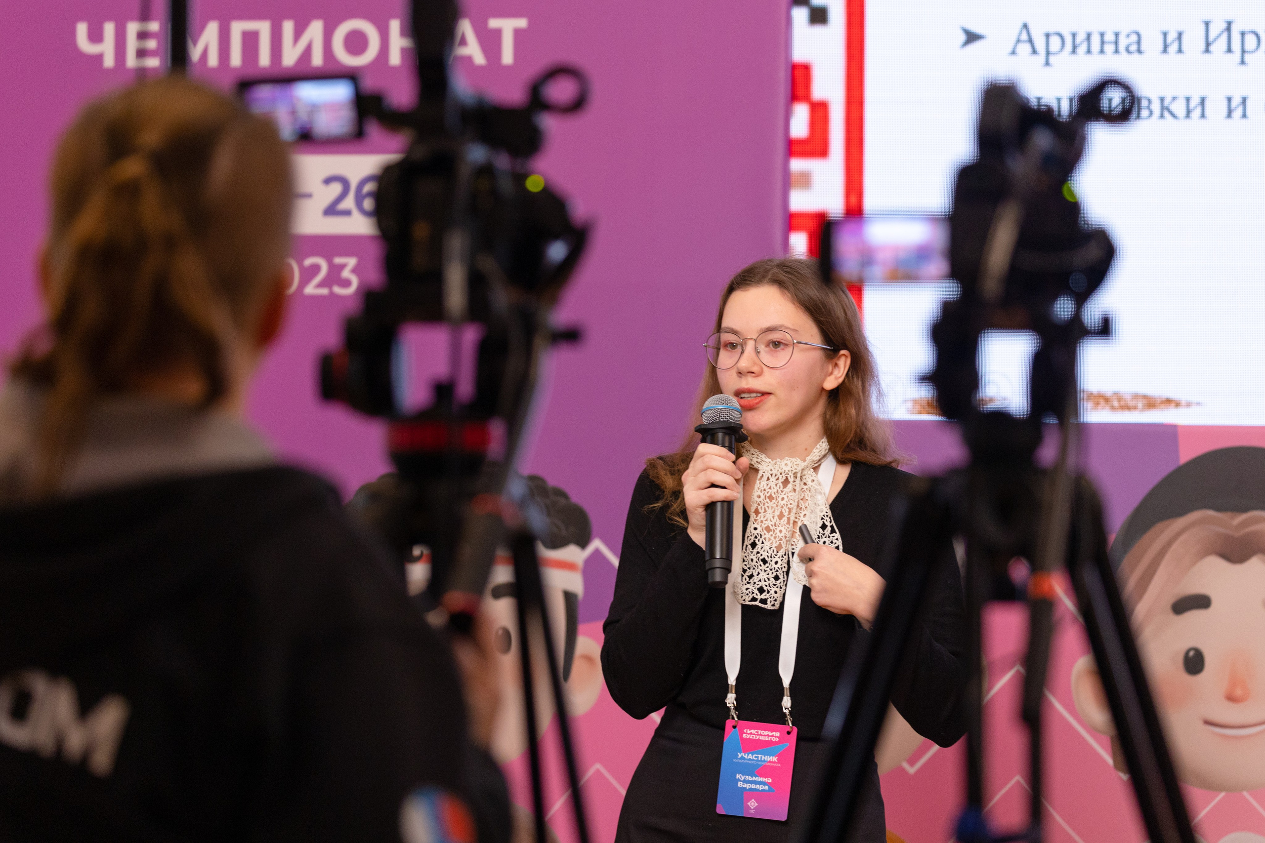 Проходит съемка выступления ведущей в очках и черной кофте.