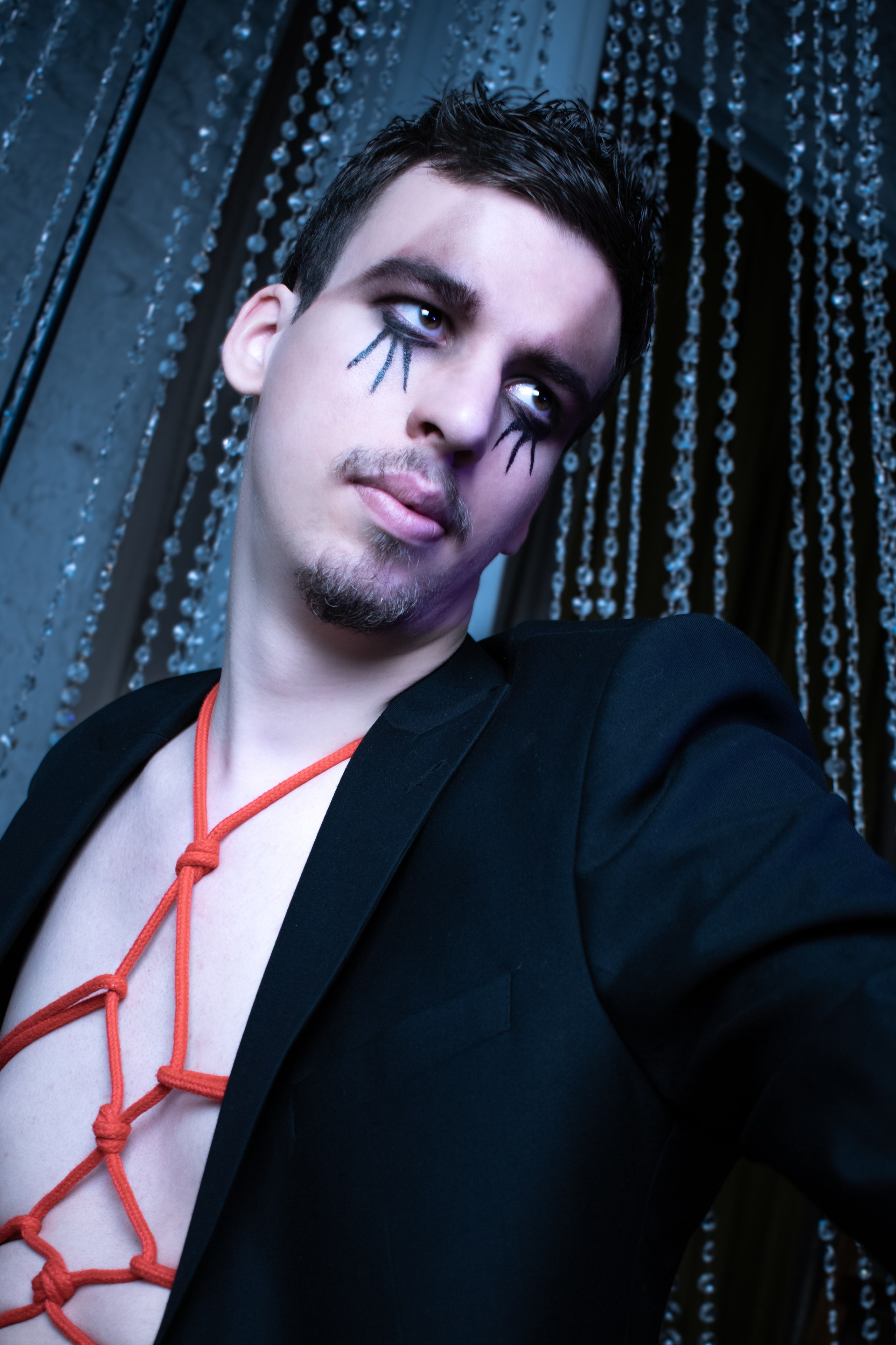 Парень в готическом макияже и черном пиджаке позирует на синем фоне