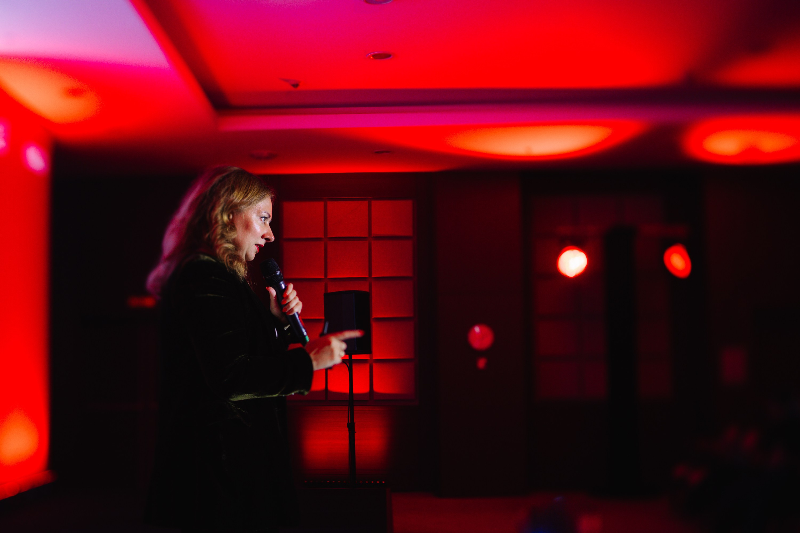 Выступление: Женщина-спикер выступает перед аудиторией в камерной обстановке с красной подсветкой.