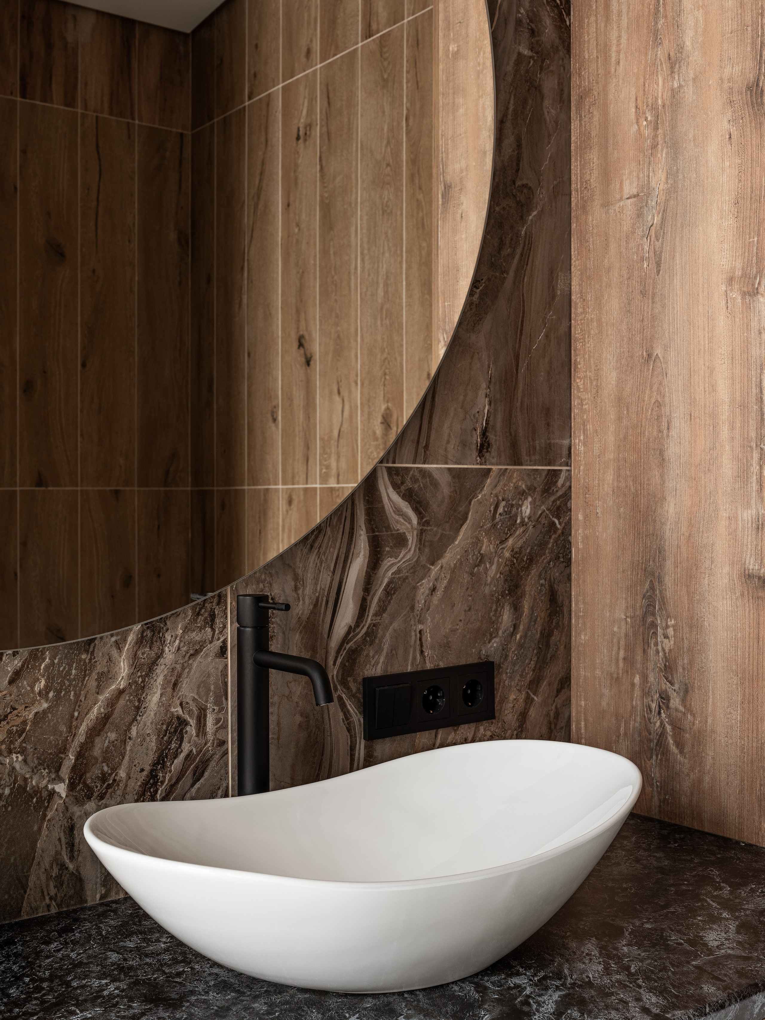 Современная белая раковина необычной формы на черной столешнице из камня, фон с отделкой деревом и мрамором, минималистичный смеситель в черном цвете; проект Екатерина Мищенко, фото Сергей Денисенко.