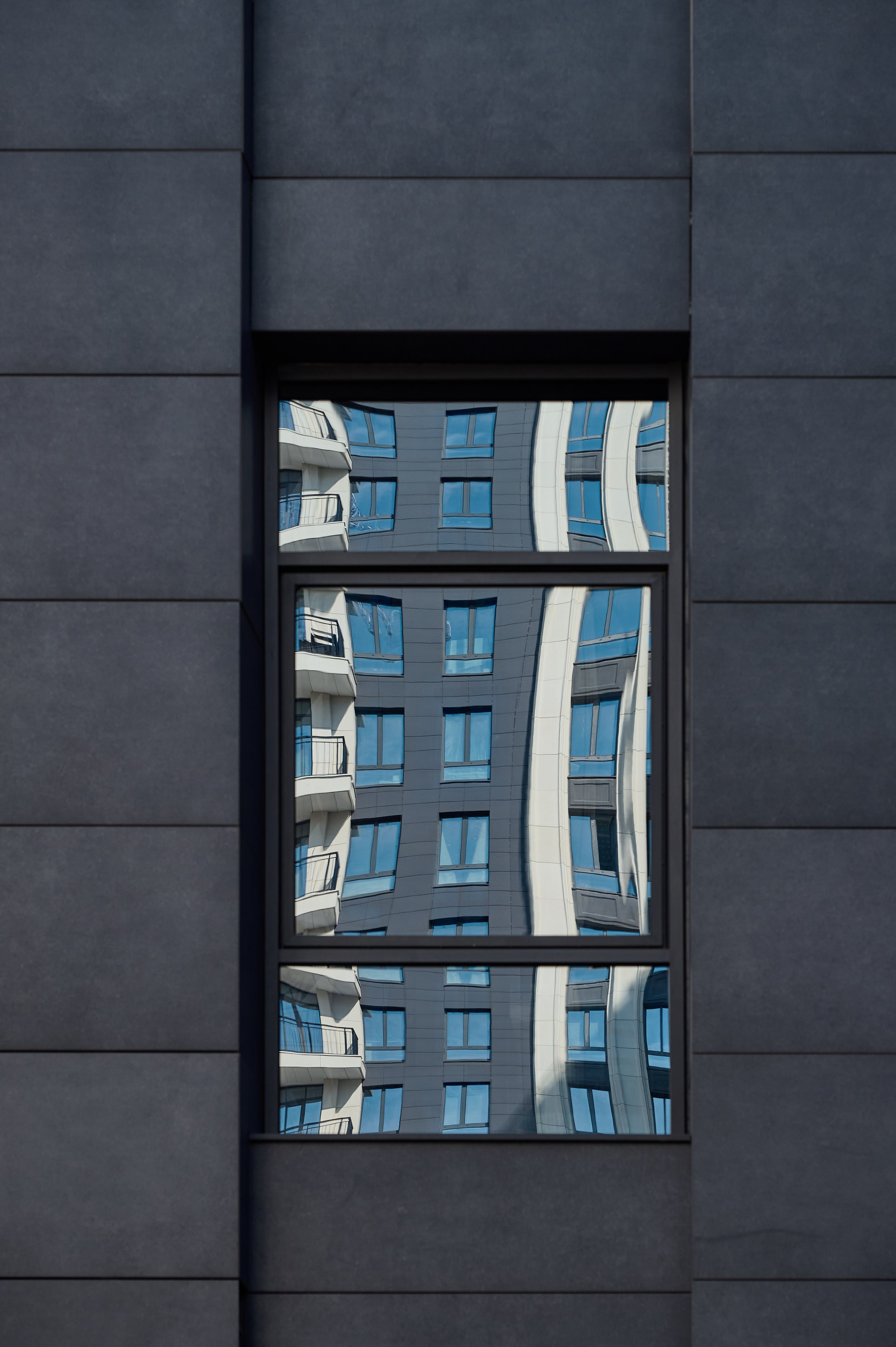 Отражение жилого здания в узком оконном проёме тёмной стены; искажённая перспектива создаёт эффект волны