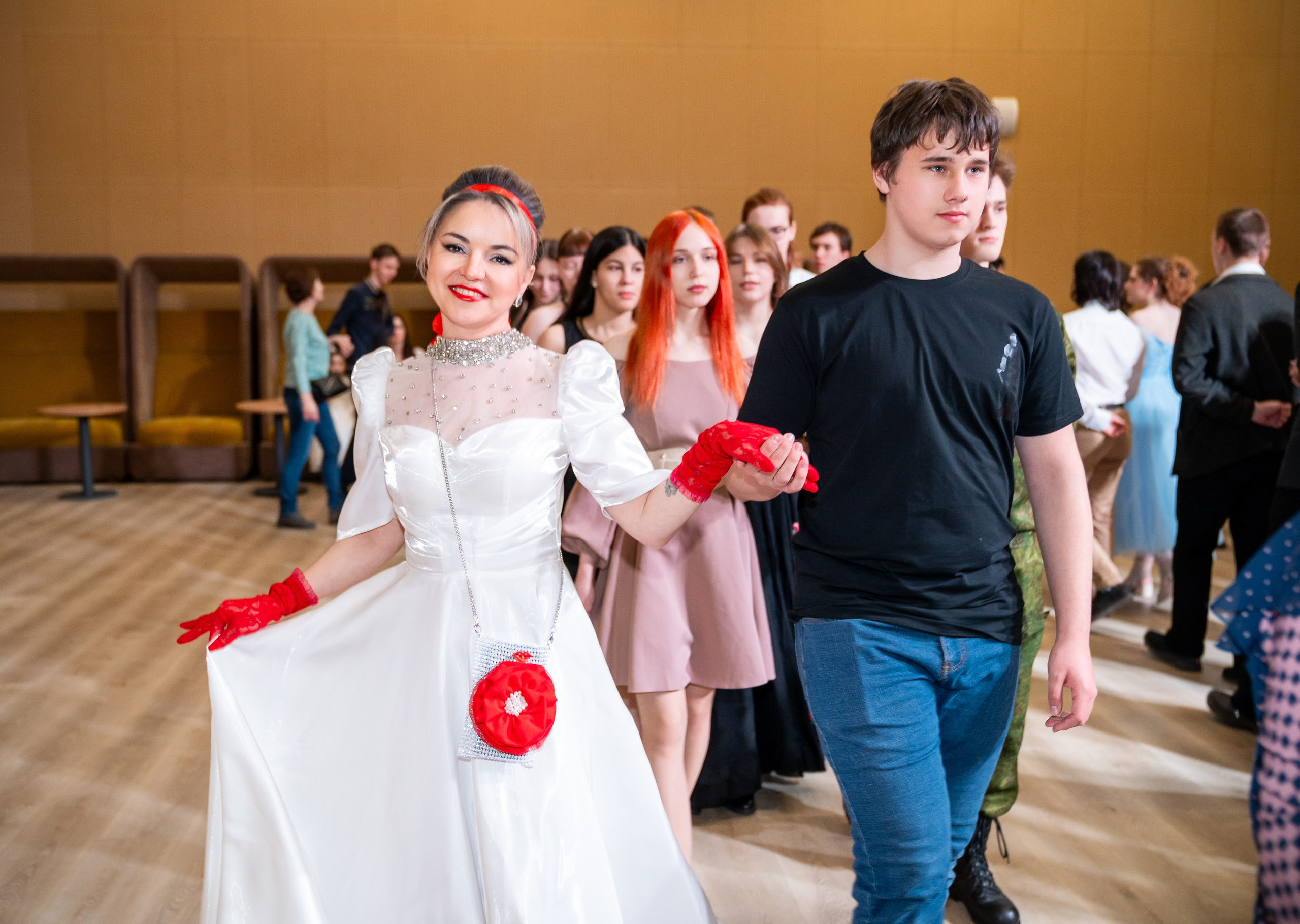 Девушка в белом платье с красными перчатками и молодой человек в чёрной футболке идут вместе на инклюзивном балу «Мы все равны». Фотограф Мальцев Евгений.