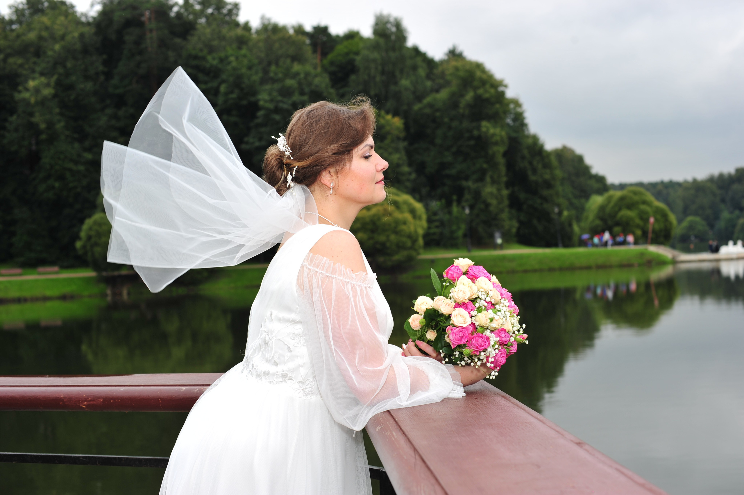 Свадьба в Царицыно пасмурным летним днем, невеста стоит, опершись на парапет, и смотрит воду пруда, короткая фата невесты развивается, в руках невеста держит свадебный букет 