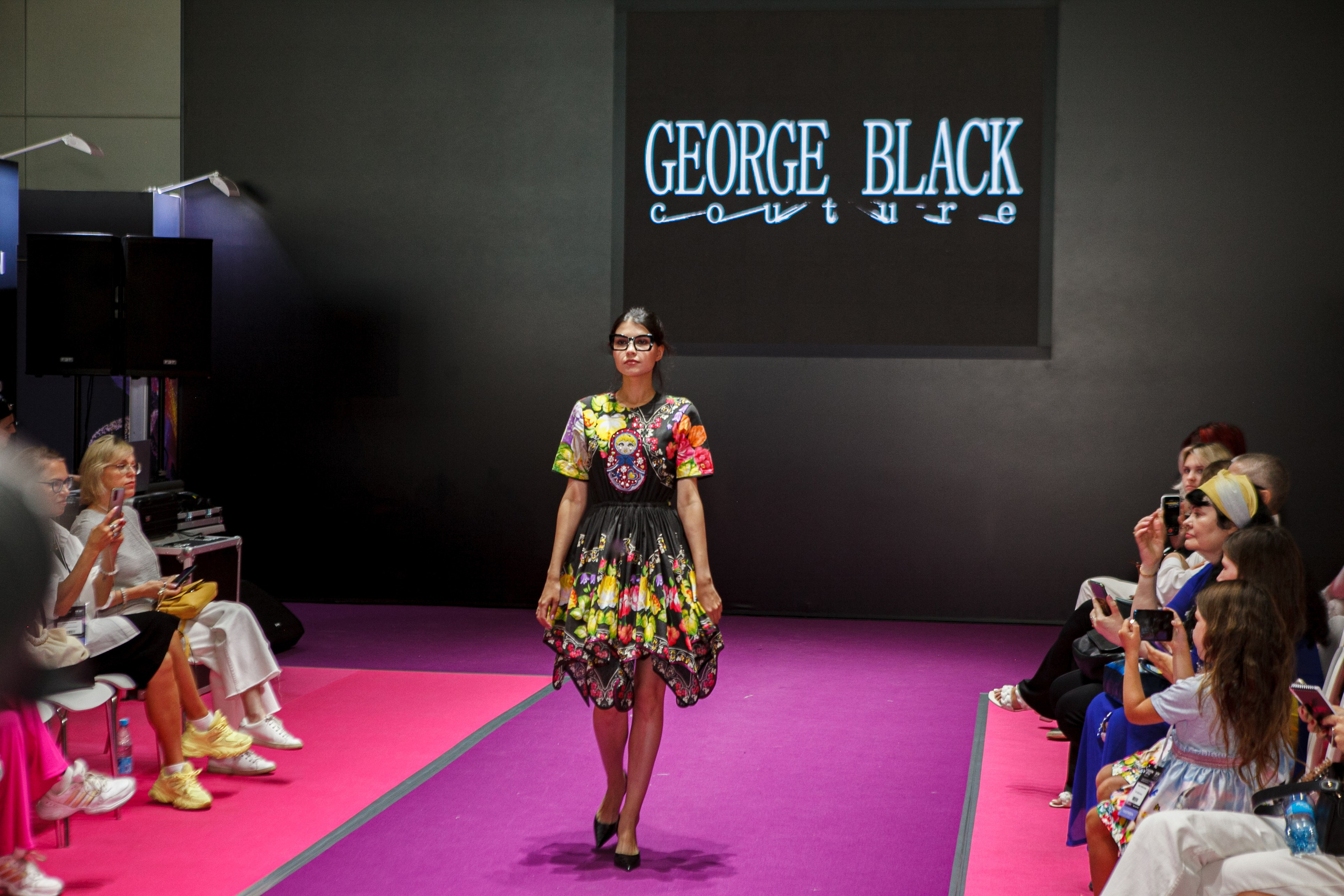  "Модели в ярких нарядах проходят по подиуму во время показа мод, освещенные яркими софитами и окруженные восторженными зрителями."