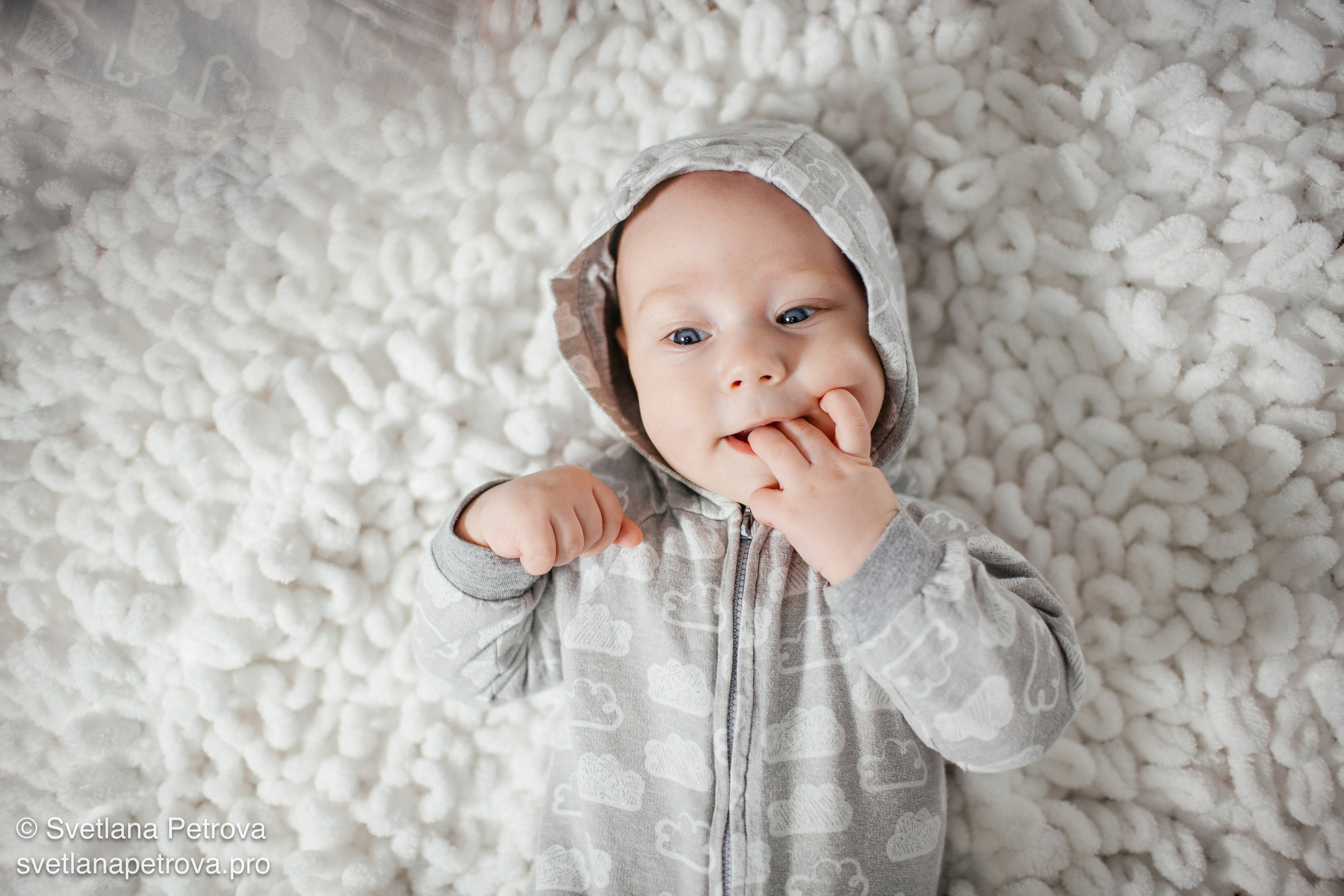 Младенец на пушистом белом покрывале, взгляд в камеру. Уютная фотосессия: малыш в сером комбинезоне. Малыш с облачками на комбинезоне — нежный кадр. Пушистый фон, младенец в капюшоне, спокойный момент. Серенький комбинезон с облаками, малыш изучает мир. Тёплый снимок: младенец на мягком покрывале. Любопытный малыш на белом ковре — эмоциональный кадр.