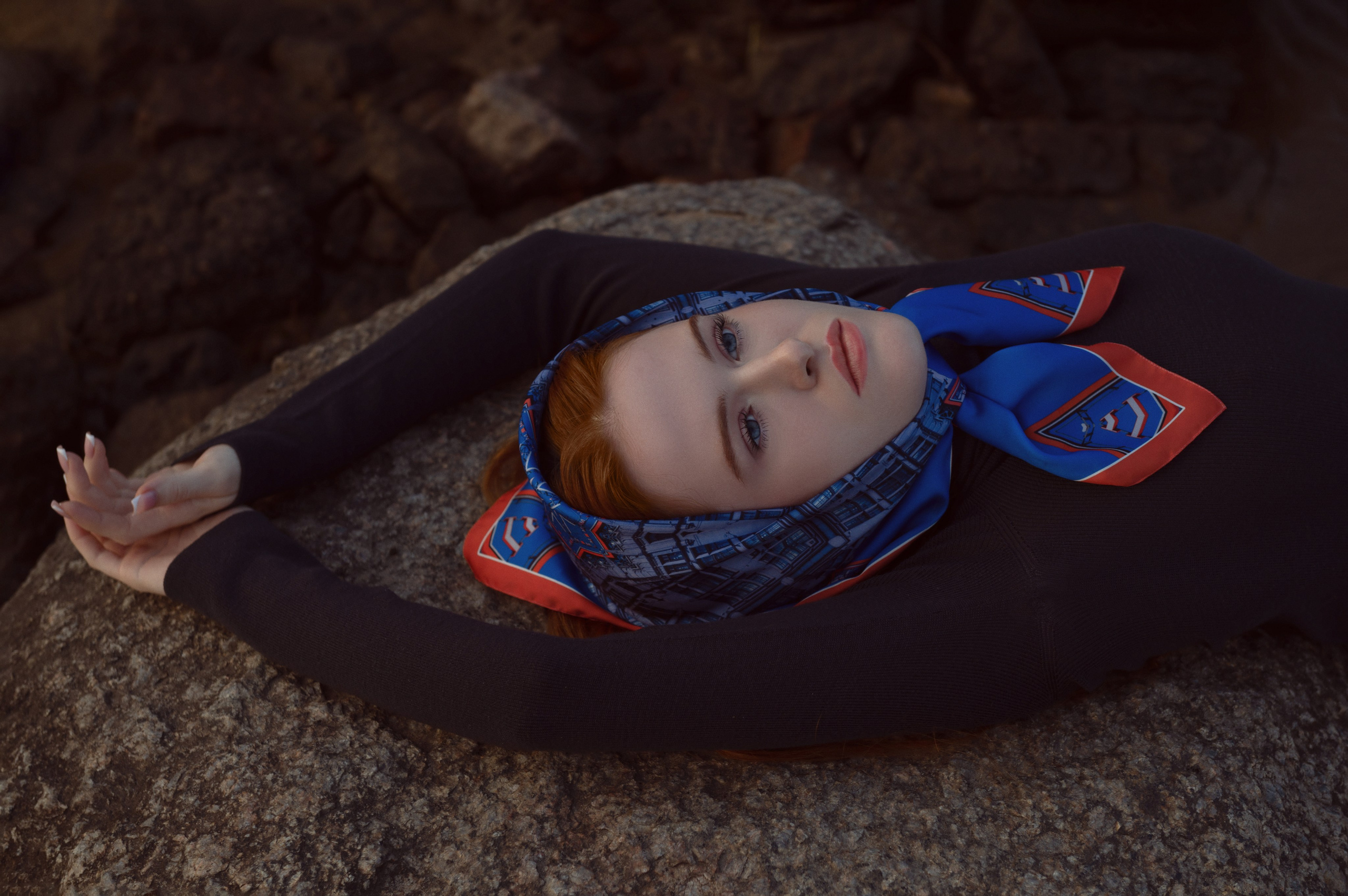 Портрет в низком ключе, красивая рыжеволосая девушка в черной водолазке лежит на сером камне, волосы ее покрывает яркий синий платок