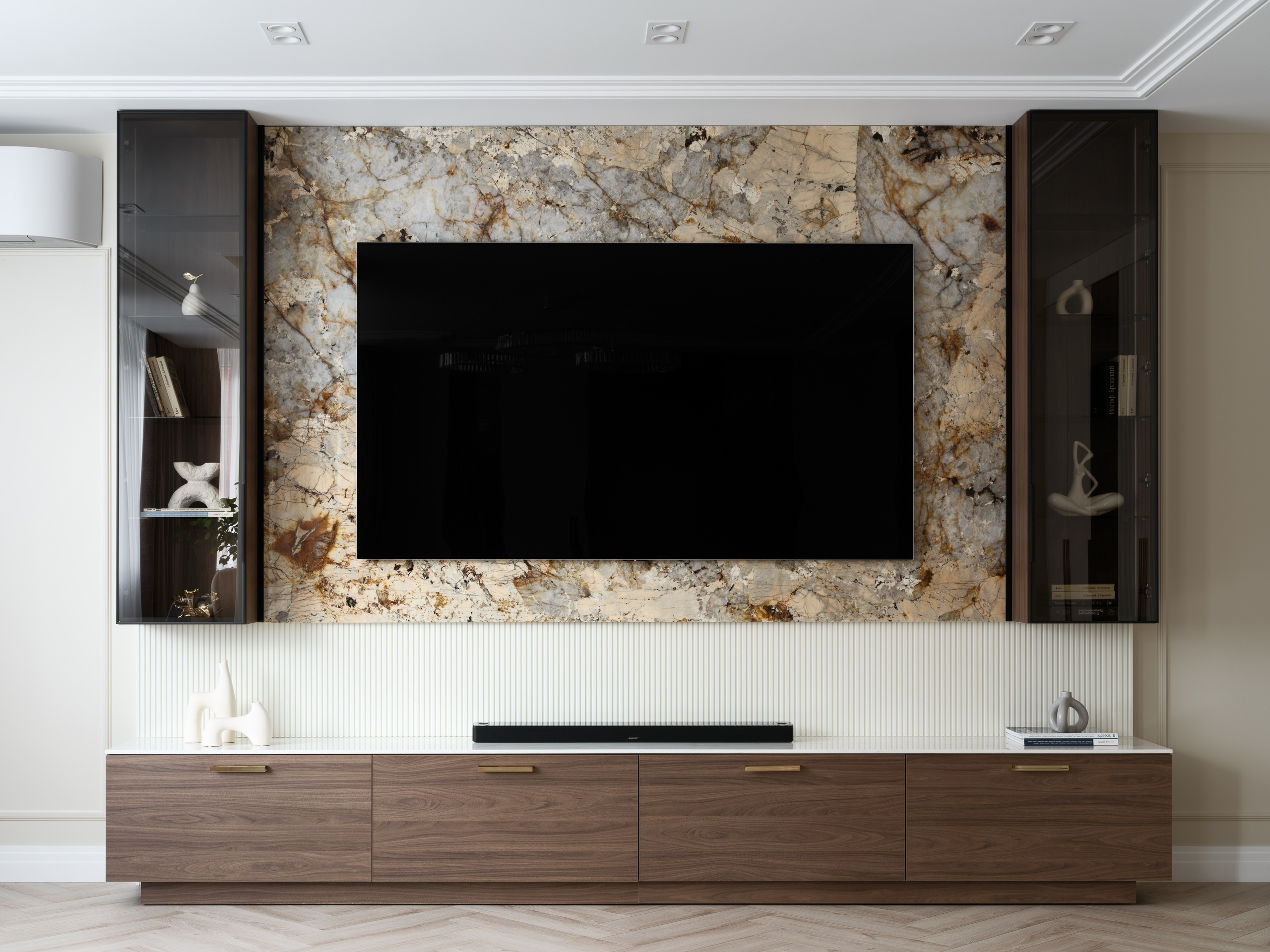 Современная гостиная с большим телевизором на фоне акцентной мраморной панели с серо-бежевыми и золотистыми прожилками, деревянными тумбами и стеклянными шкафами по бокам, выполненная в стиле современная классика.