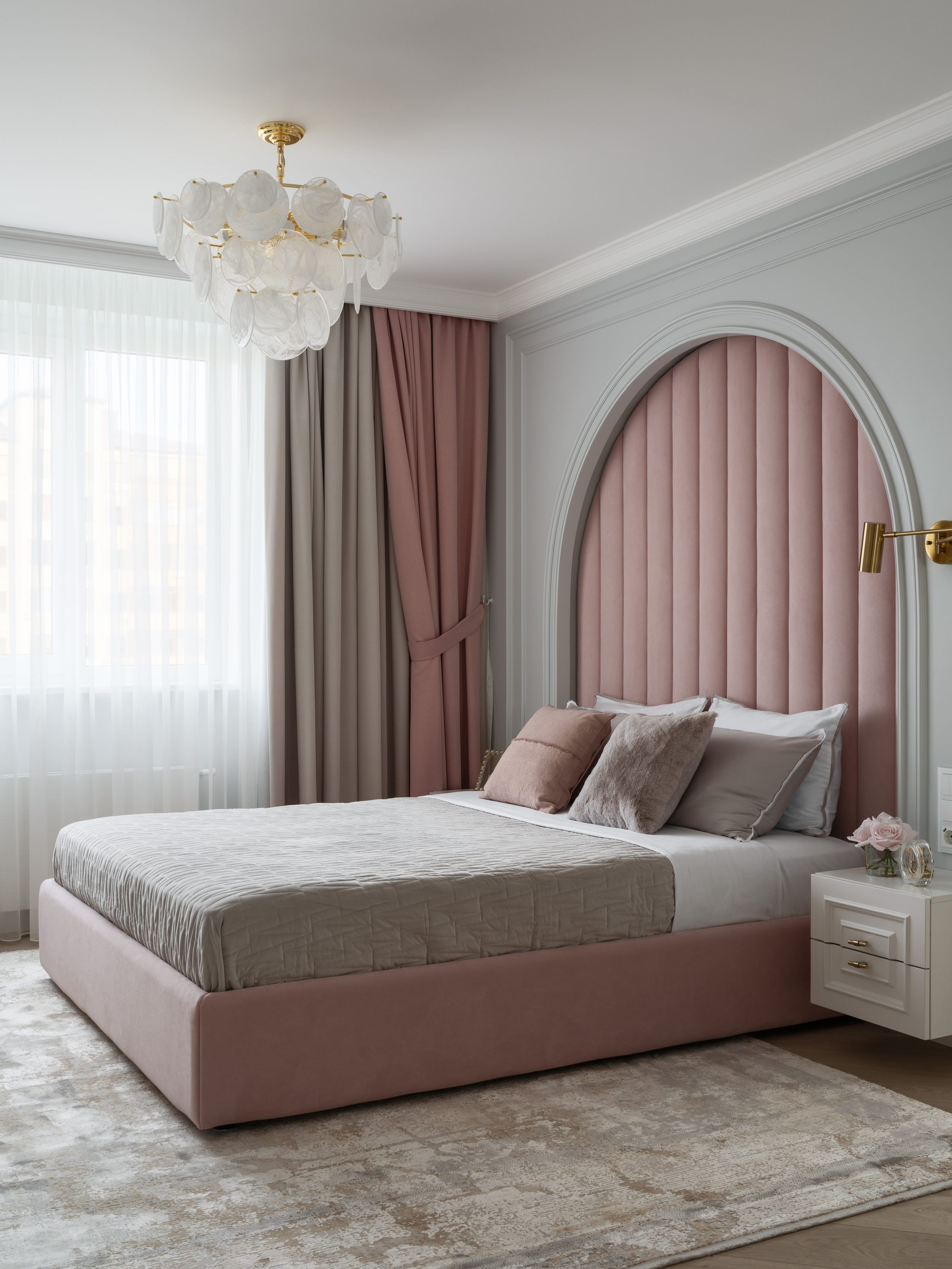 Современная спальня с мягким изголовьем арочной формы в розовом велюре, белыми тумбами и золотыми бра. Элегантный светильник завершает образ. Дизайн Евгении Ермошиной, фото Сергея Денисенко.