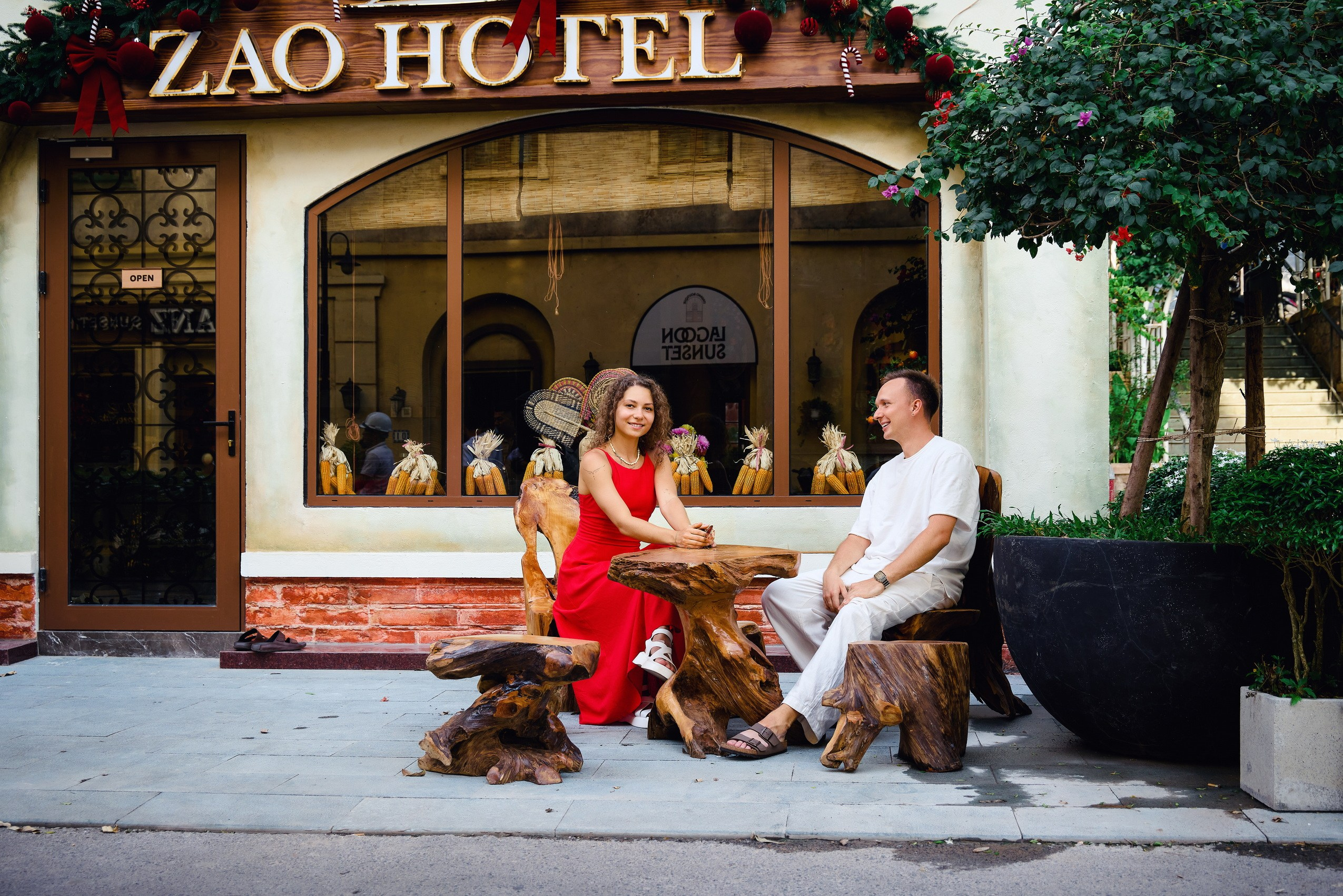 Couple in red and white clothes sitting on natural wooden stools outside ZAO Hotel, a building with Mediterranean-style architecture in Sunset Town, Phu Quoc.