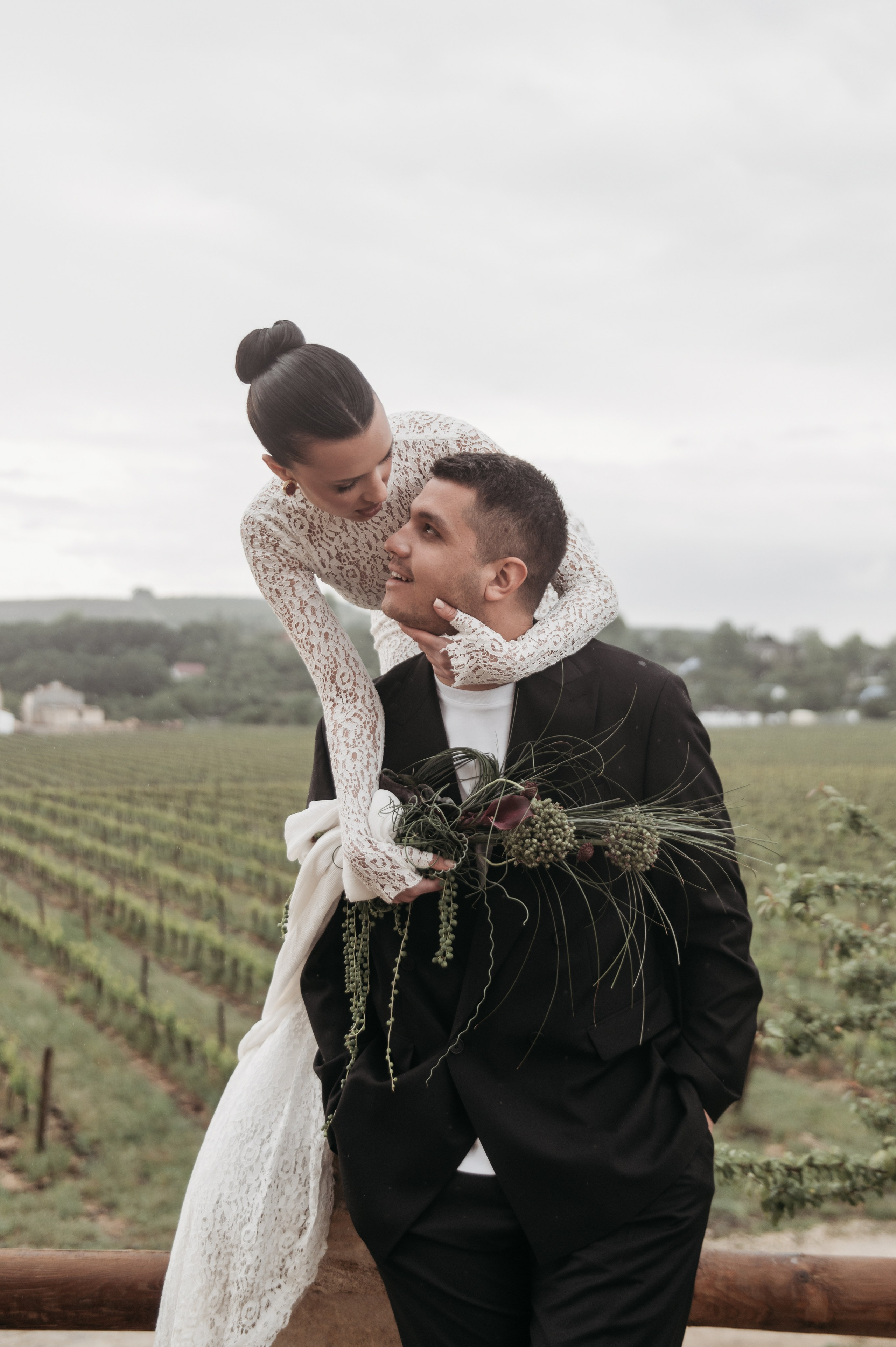 Невеста в кружевном свадебном платье обнимает жениха, который смеется, на фоне виноградника, создавая атмосферу счастья и романтики