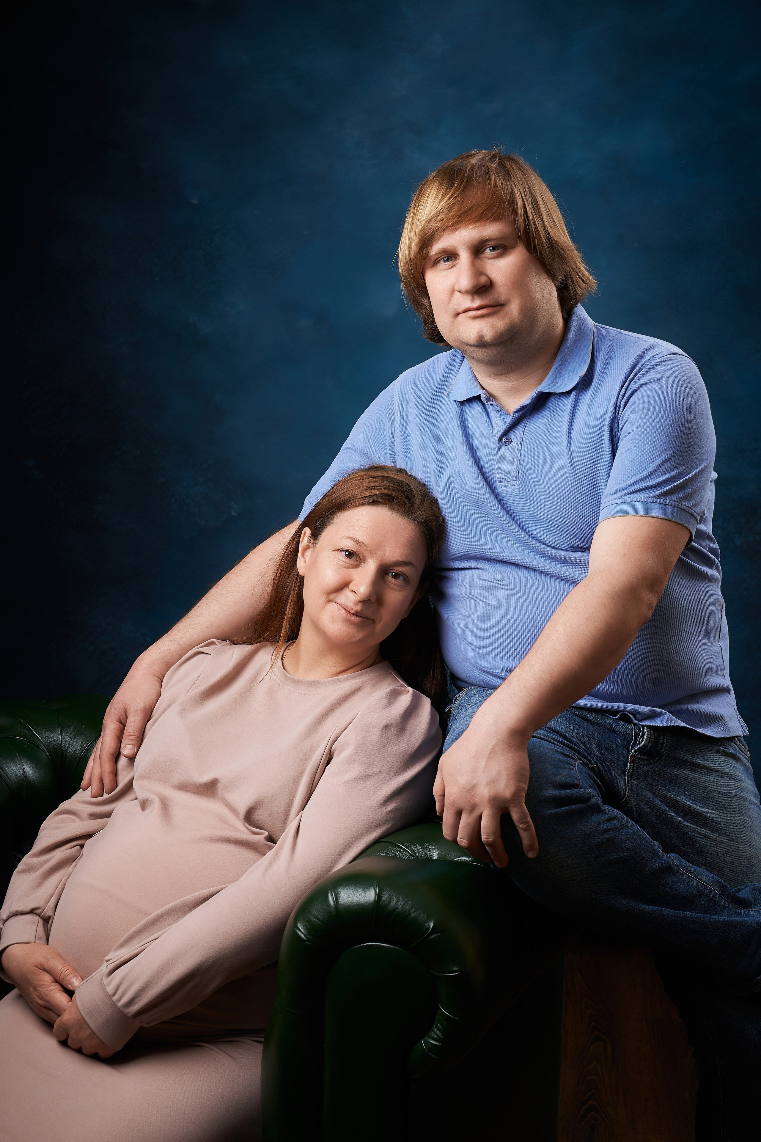 беременная жена сидит на зелёном кожаном кресле и её обнимет муж на семейной фотосессии