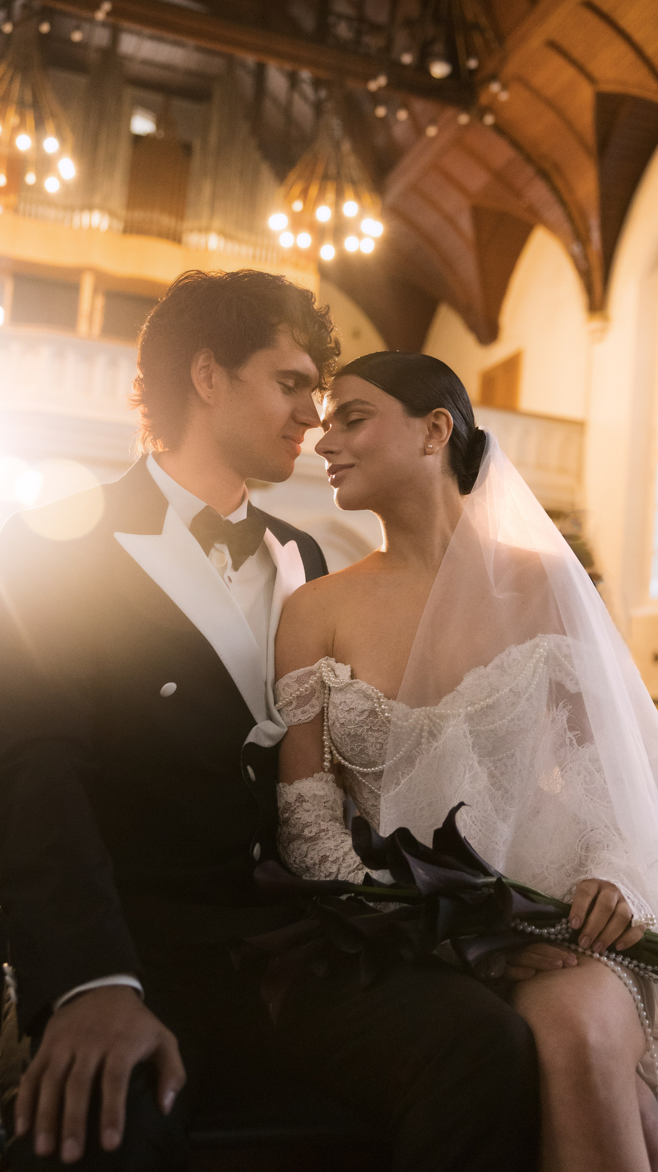Романтический свадебный портрет жениха и невесты крупным планом, сидящих рядом в тёплом, сияющем интерьерном свете. Невеста одета в платье с открытыми плечами из жемчужного кружева и держит в руках тёмный букет калл, а жених одет в чёрный смокинг с белыми лацканами.