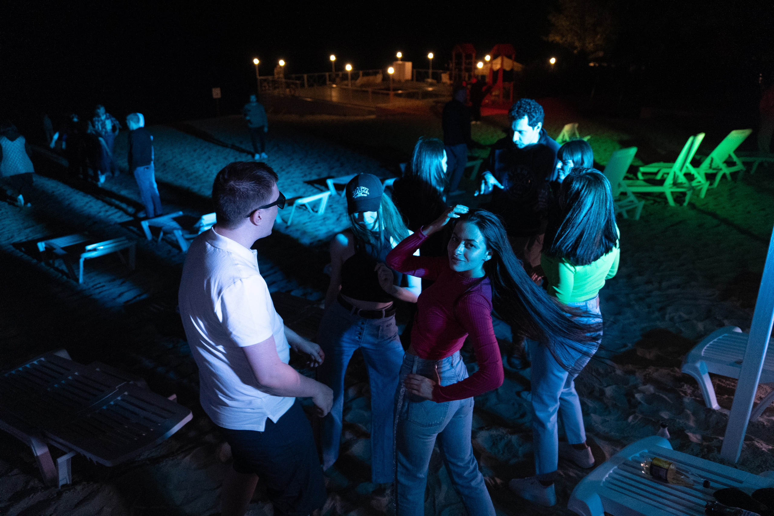 Молодежная вечеринка на пляже ночью, группа друзей танцует на песке, светлые лежаки