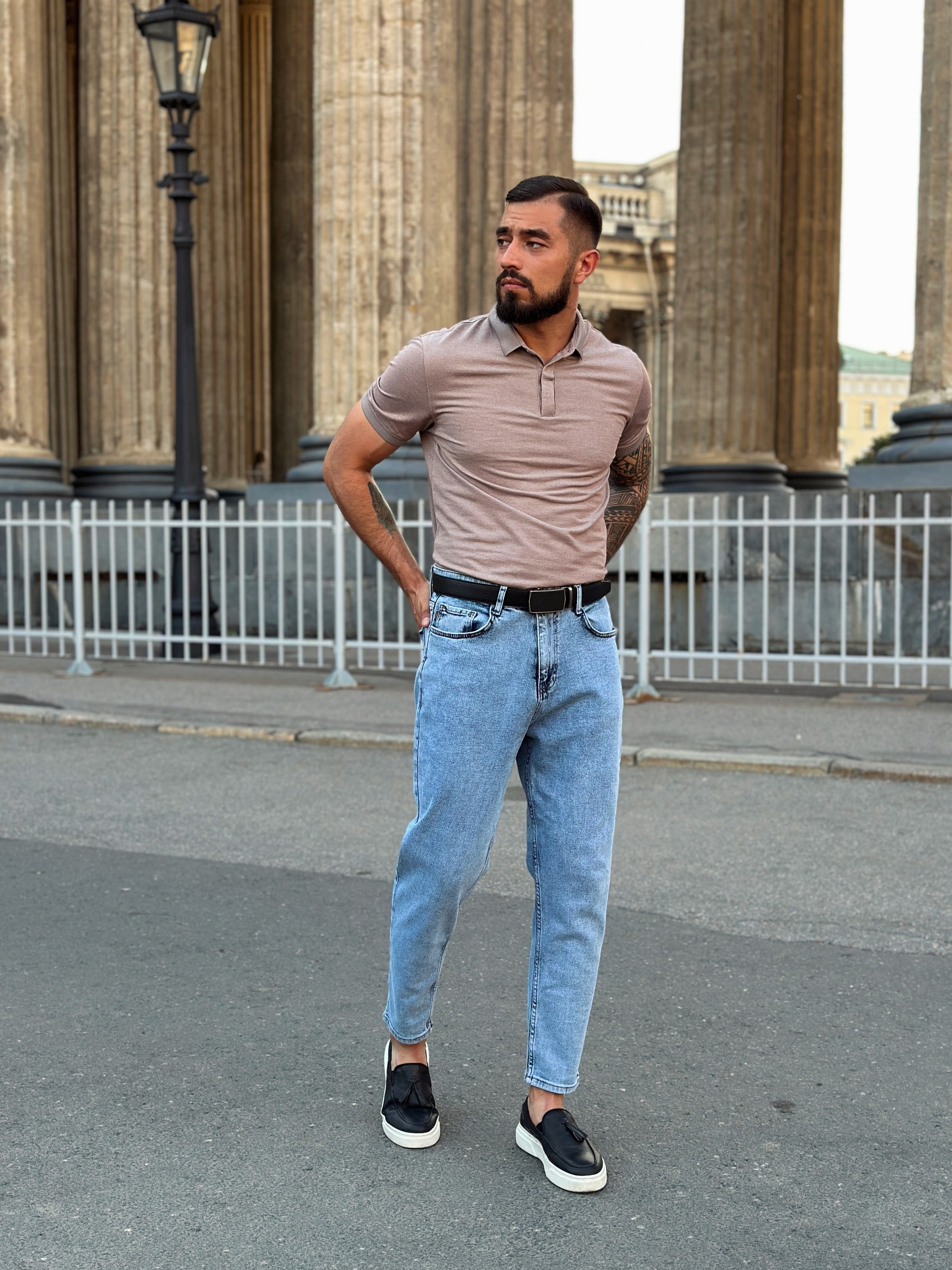 Мужчина модель с бородой в синих джинсах и поло идет возле Казанского собора в СПб