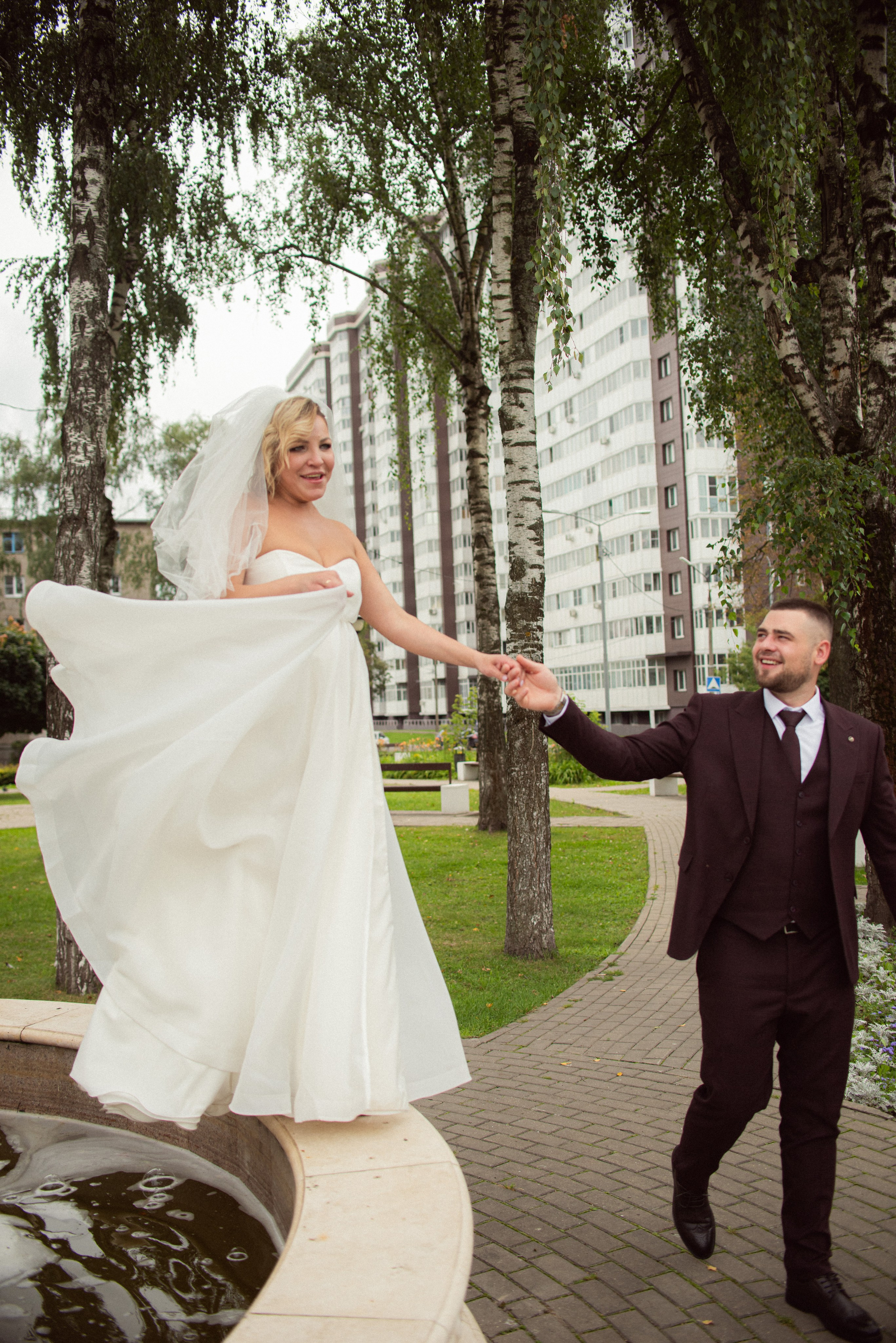 Невеста в свадебном платье гуляет по краю фонтана, жених рядом, фотосессия в Купавне