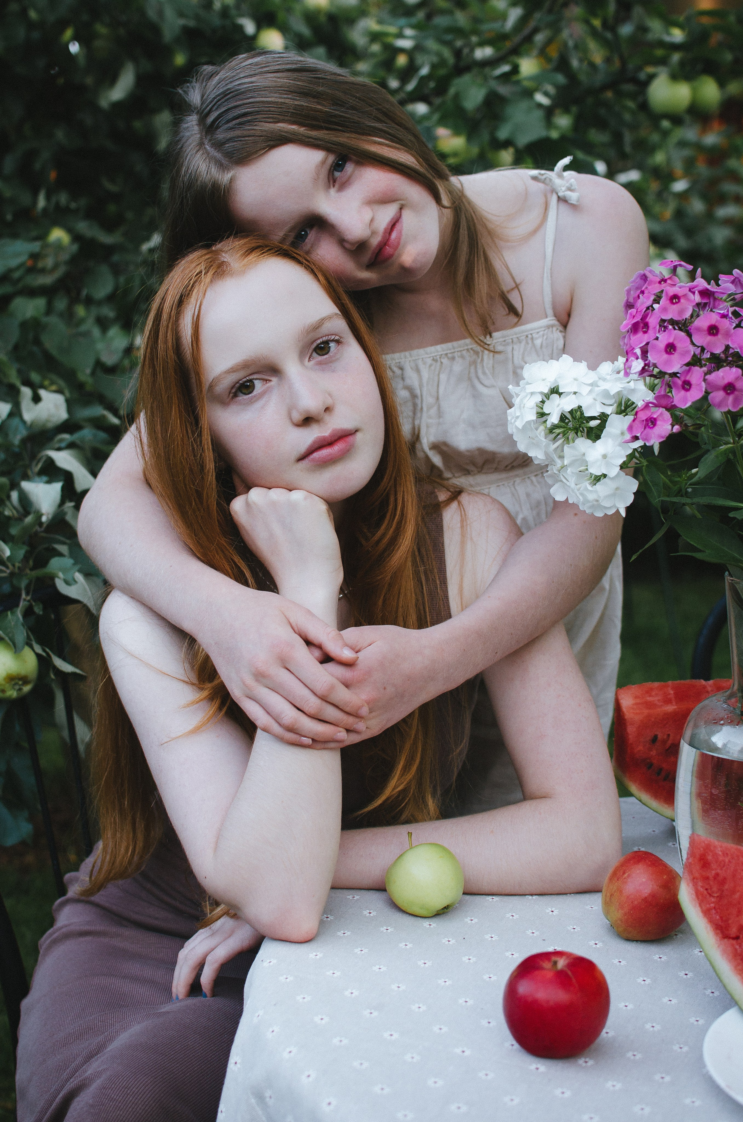 Две сестры позируют в саду за столом с красными яблоками.