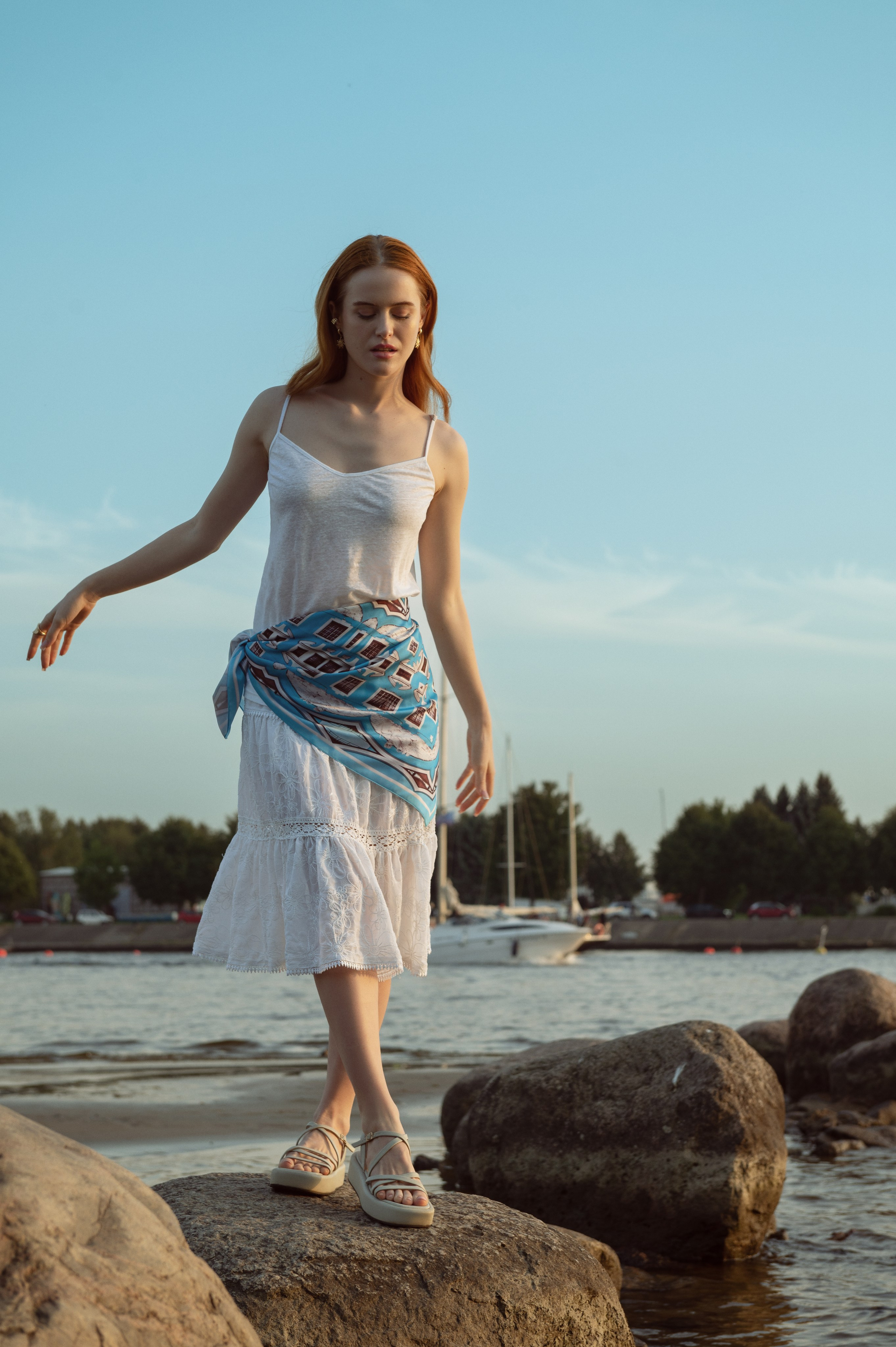 Красивая рыжеволосая девушка в белом топе и белой юбке стоит на большом камне, на бедрах у нее повязан голубой платок. На заднем плане река и белоснежные яхты.