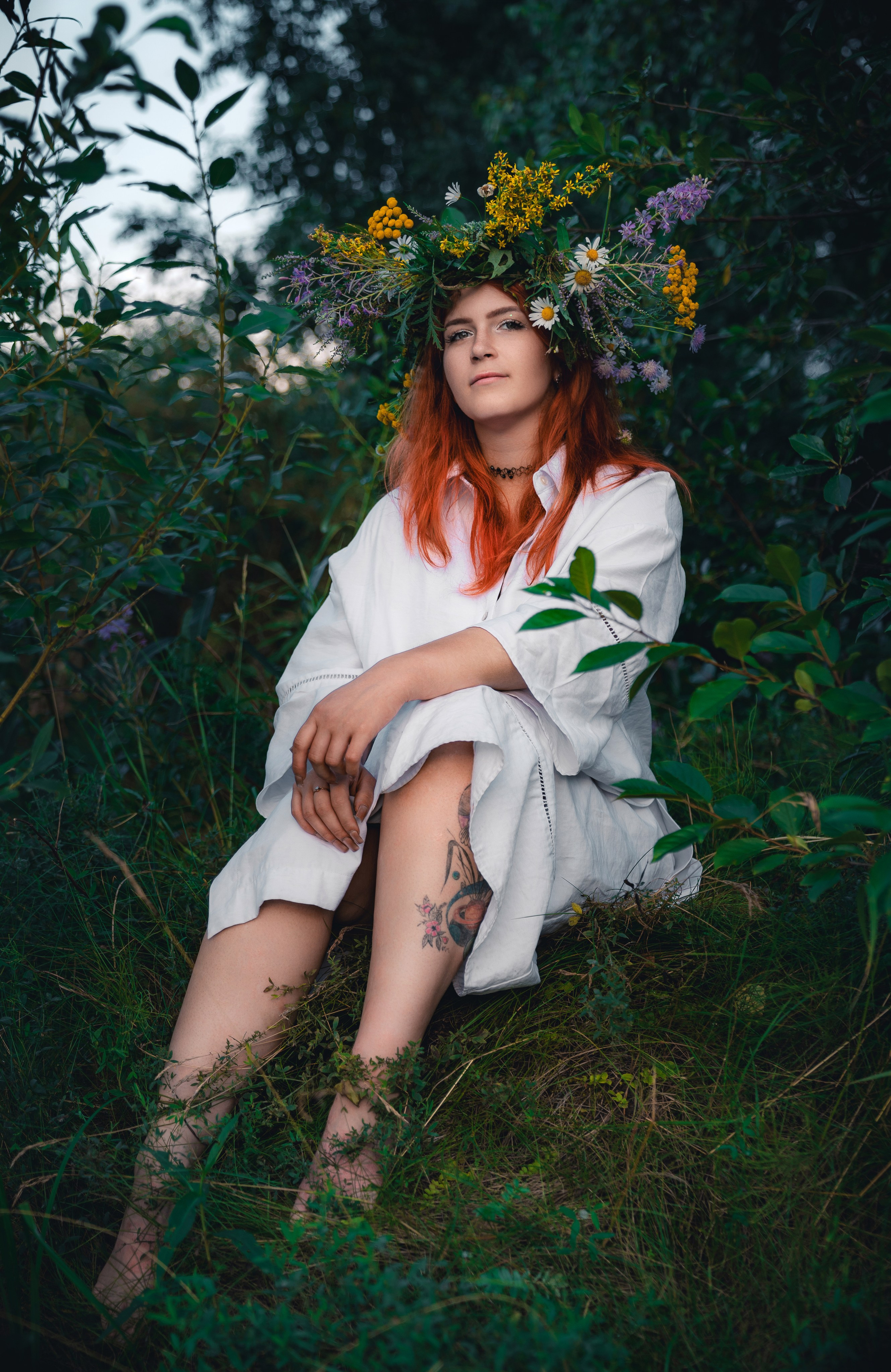 Фотосессия с венком из полевых цветов: рыжеволосая девушка сидит в траве, татуировка на ноге, естественный портрет на закате.
