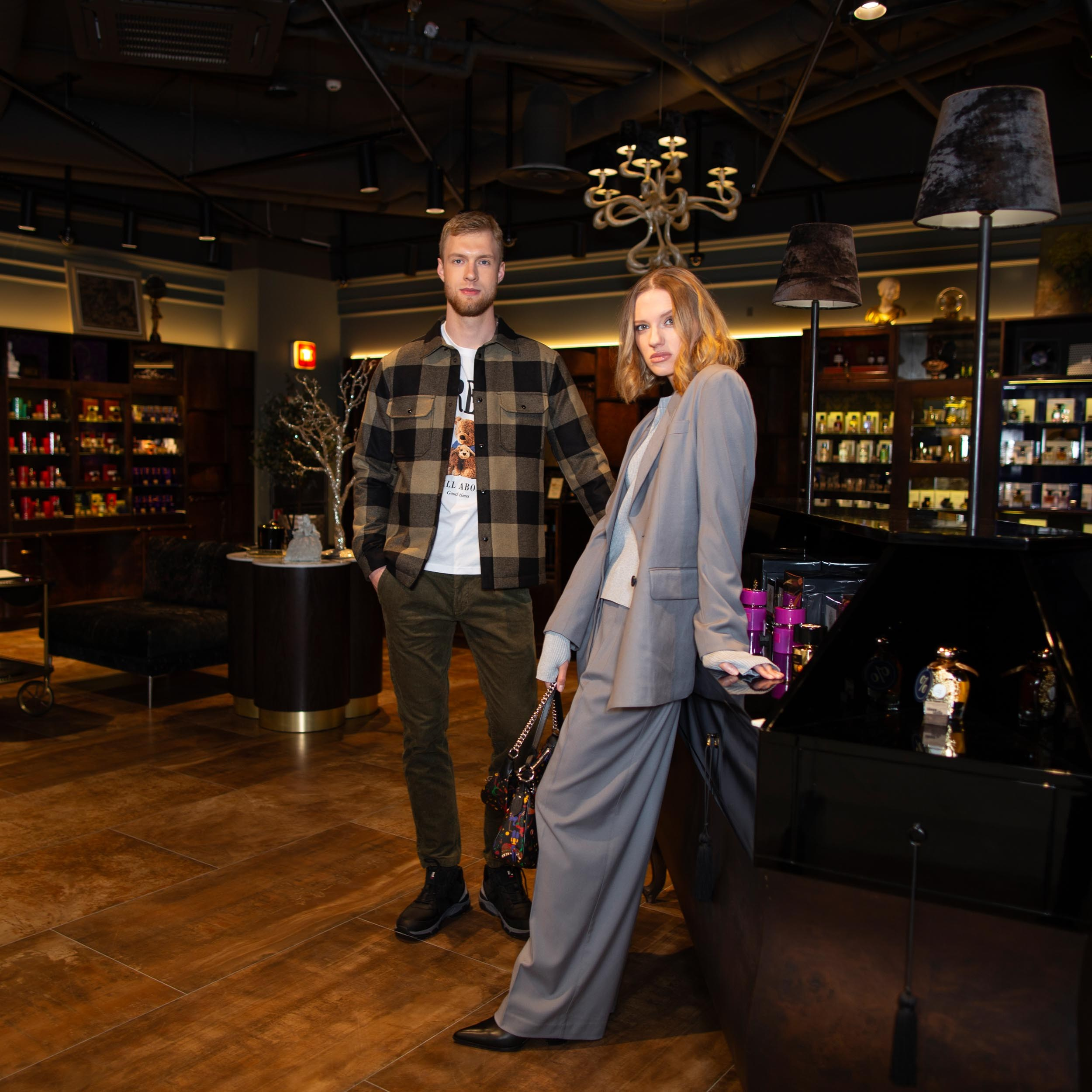 Парень с бородой в клетчатой куртке-рубашке и девушка в брючном костюме стоят в интерьере магазина