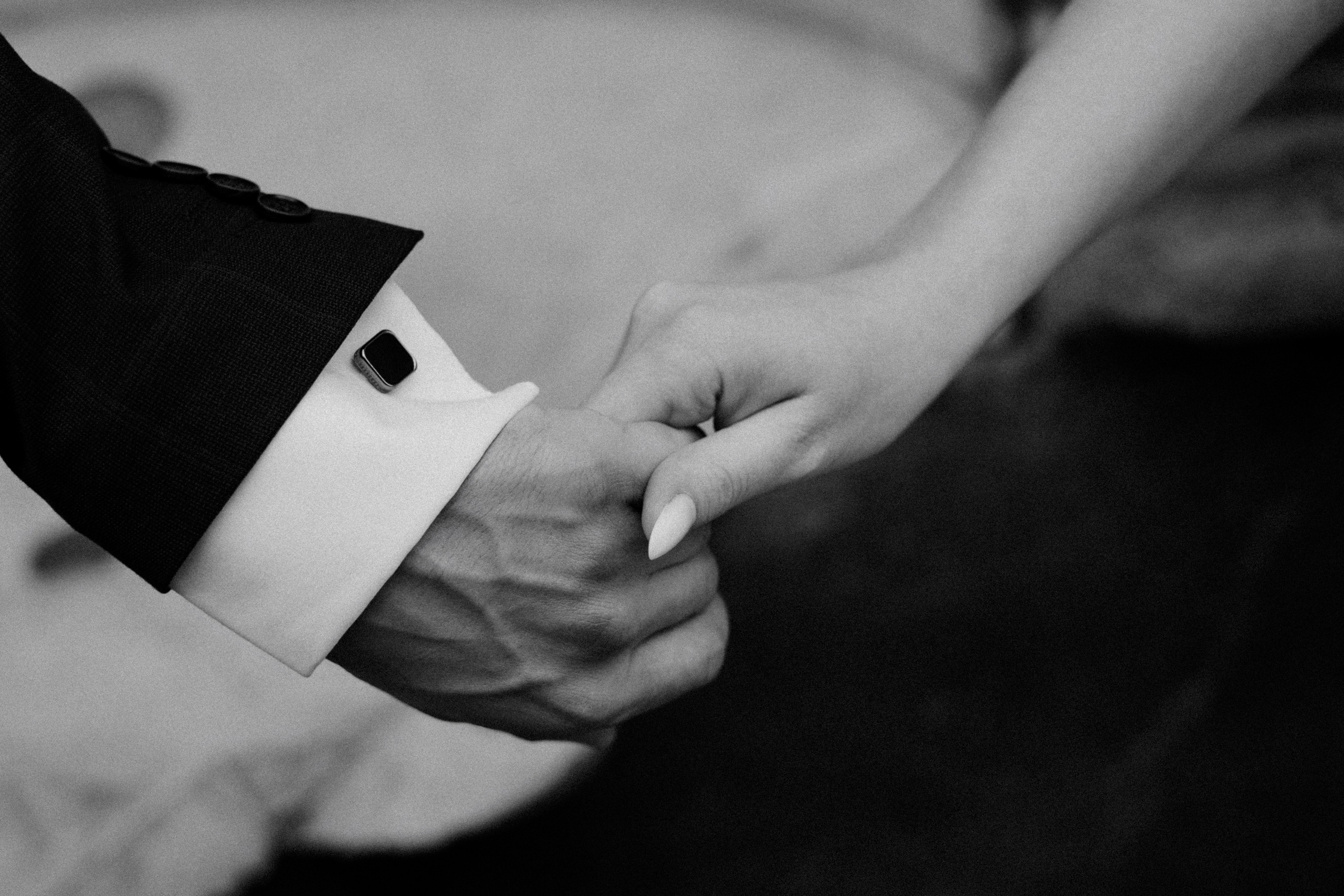 Черно-белое фото: крупный план рук жениха и невесты, держащихся друг за друга. Детали свадебной церемонии, кольца на пальцах.