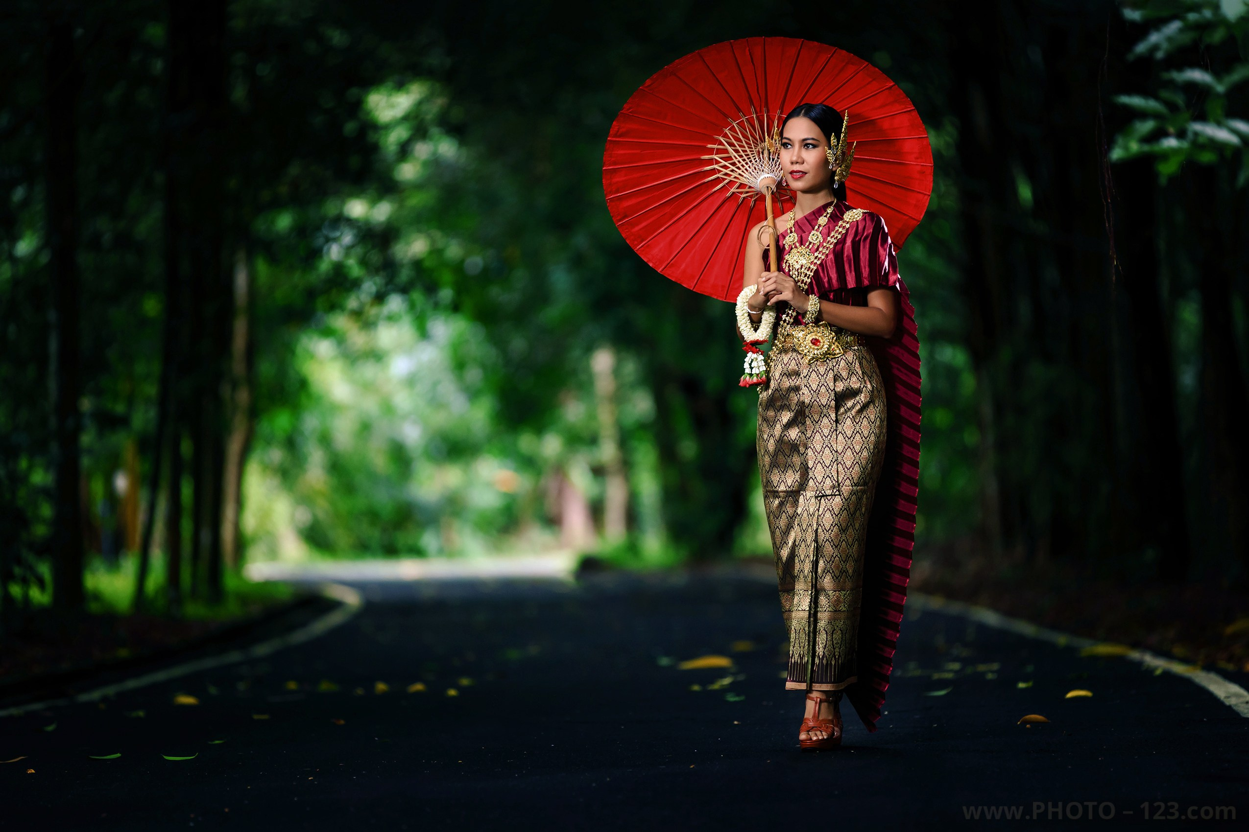 Portrait of a beautiful Thai woman wearing traditional Thai costume and holding a red parasol, walking along a forest road, photographed by a commercial photographer in Phuket