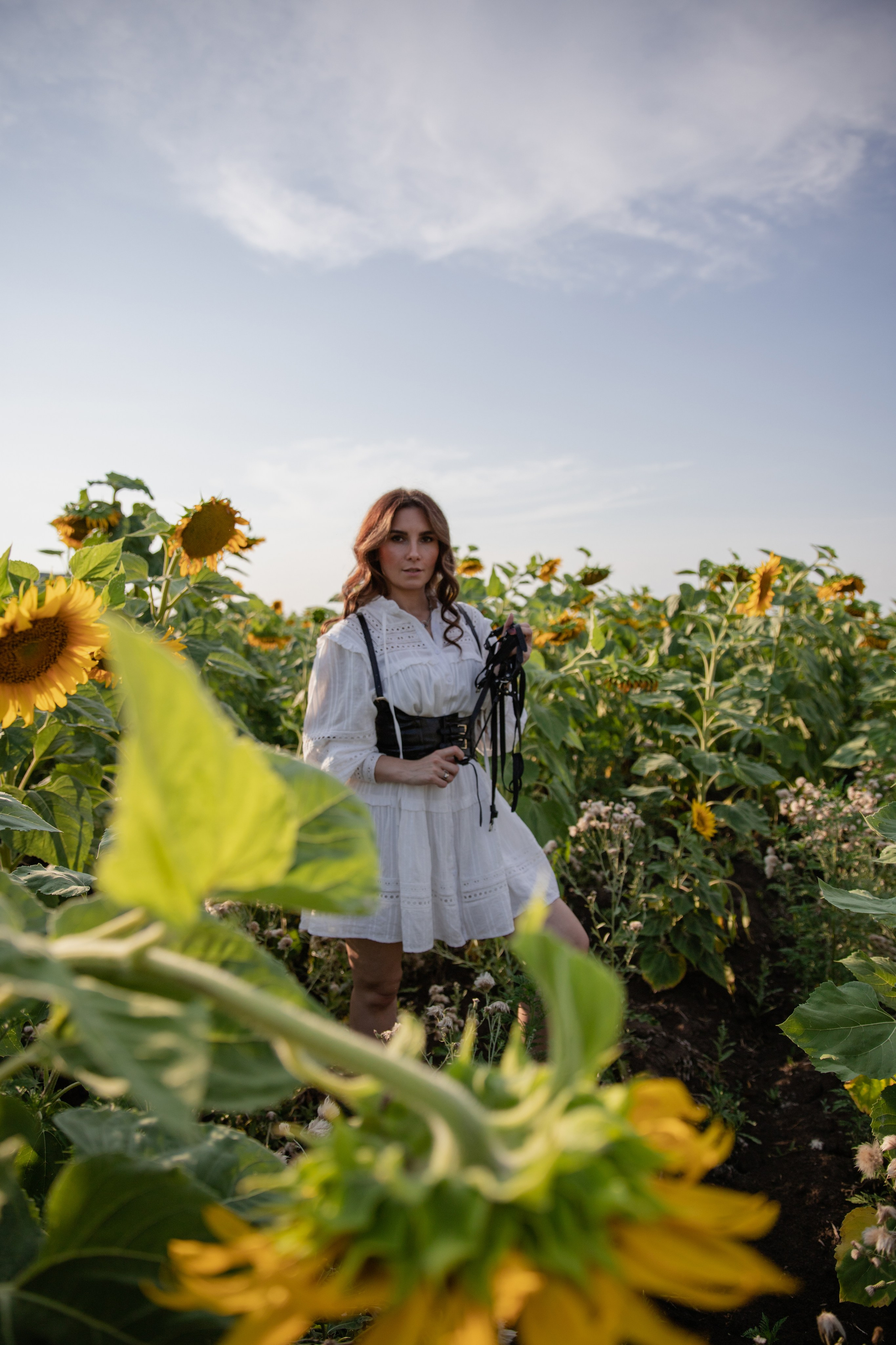 Девушка в  в белом платье  с портупеей, стоящая среди ярких подсолнухов, улыбающаяся к камере на закате