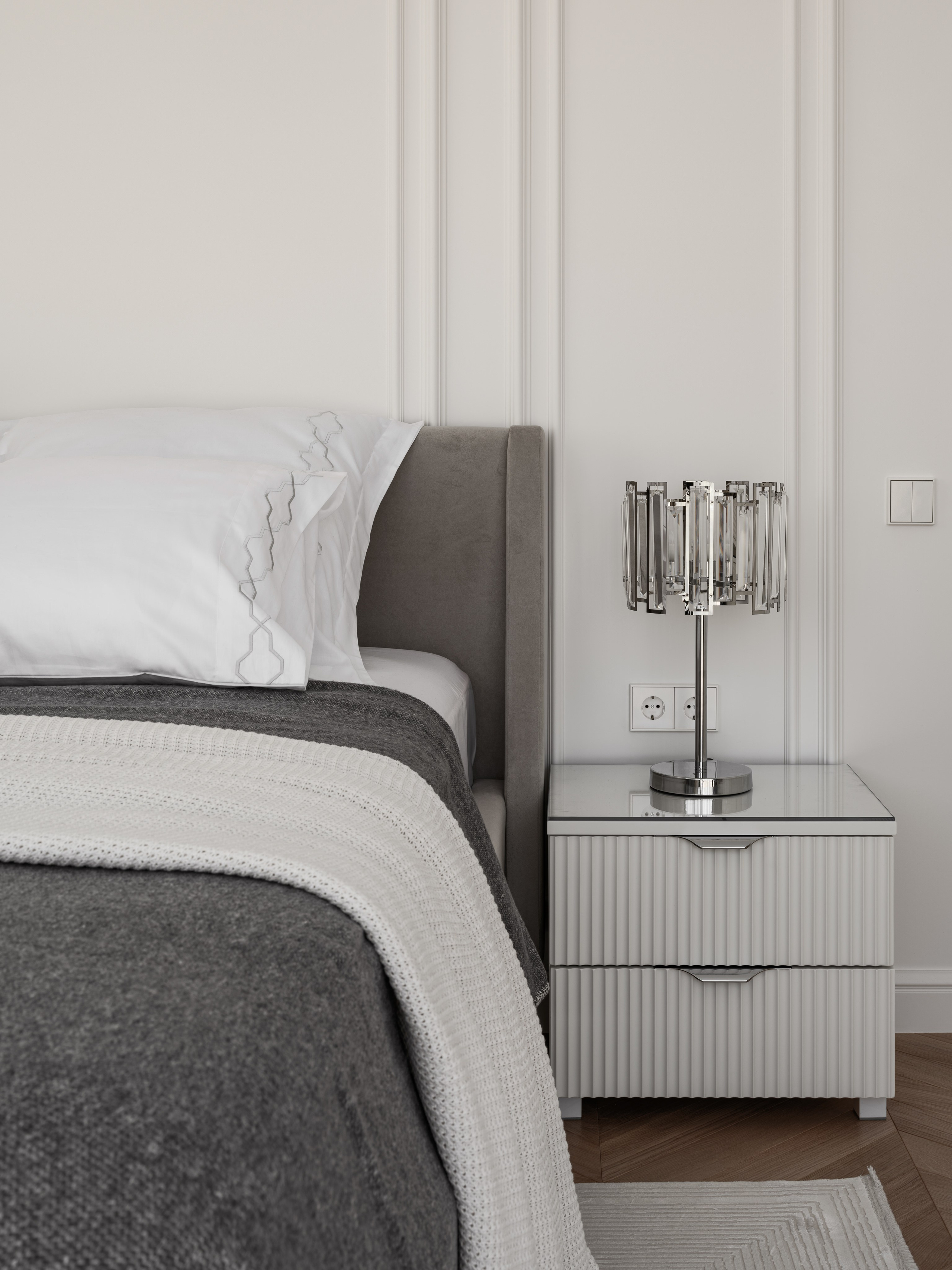 Элегантная прикроватная зона спальни с белой тумбой, стеклянной лампой и мягким текстилем в оттенках серого и молочного, разработанная дизайнером Алёной Боярской. Стилист Наталья Якушева добавила утончённые детали, фотограф Сергей Денисенко передал мягкость света и текстур.