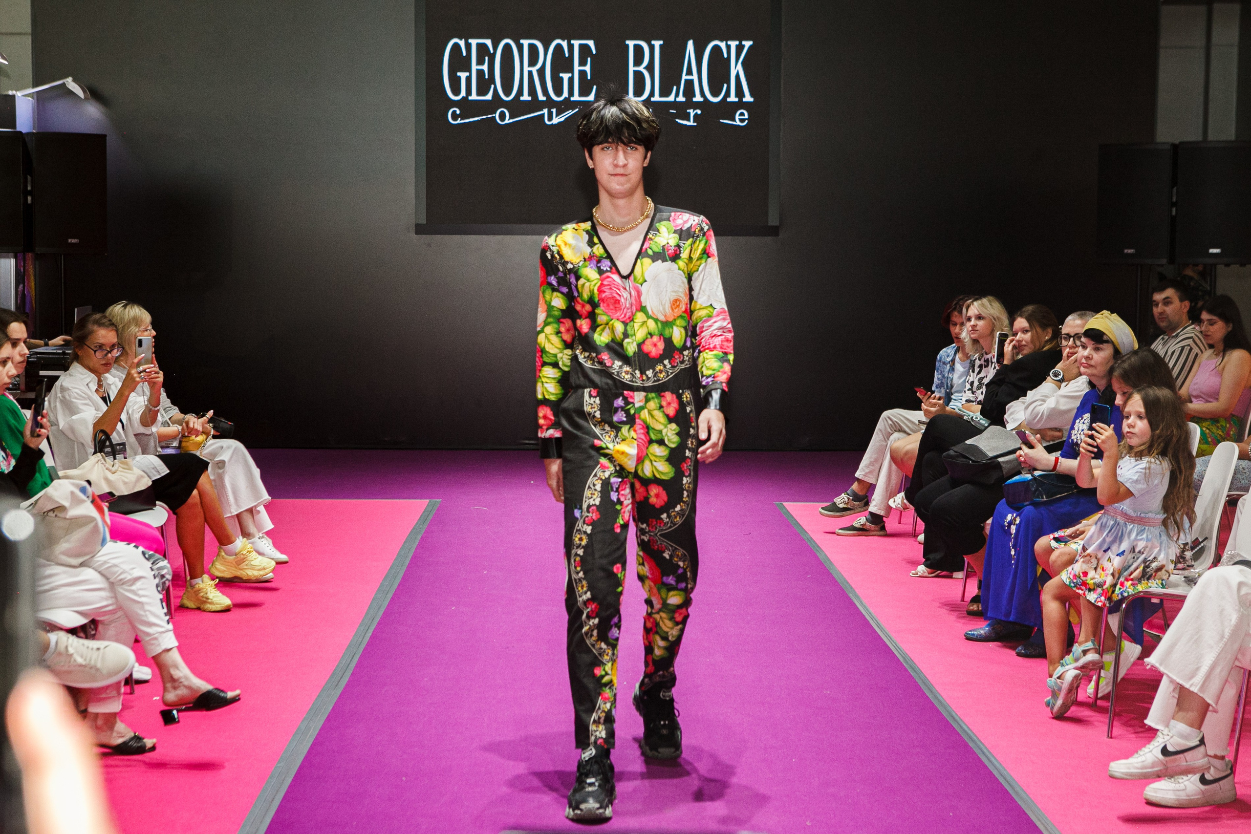  "Модели в ярких нарядах проходят по подиуму во время показа мод, освещенные яркими софитами и окруженные восторженными зрителями."