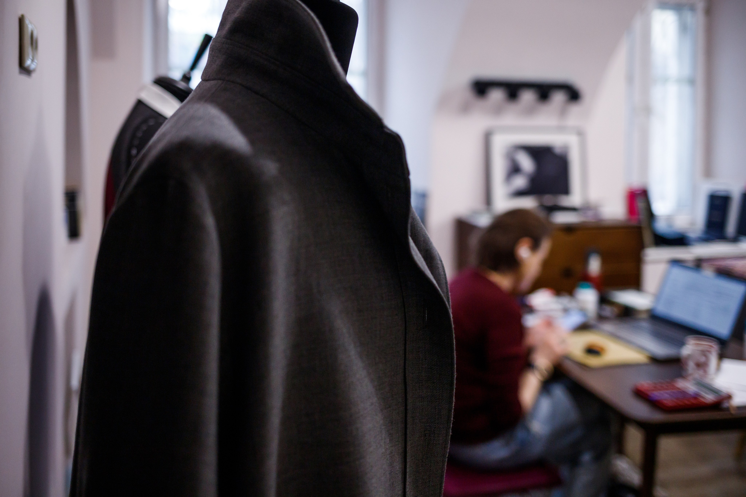 Серый пиджак на манекене, мастер за рабочим столом на втором плане, ателье Natalia Chevdar, Екатеринбург, фотограф Илья Антропов