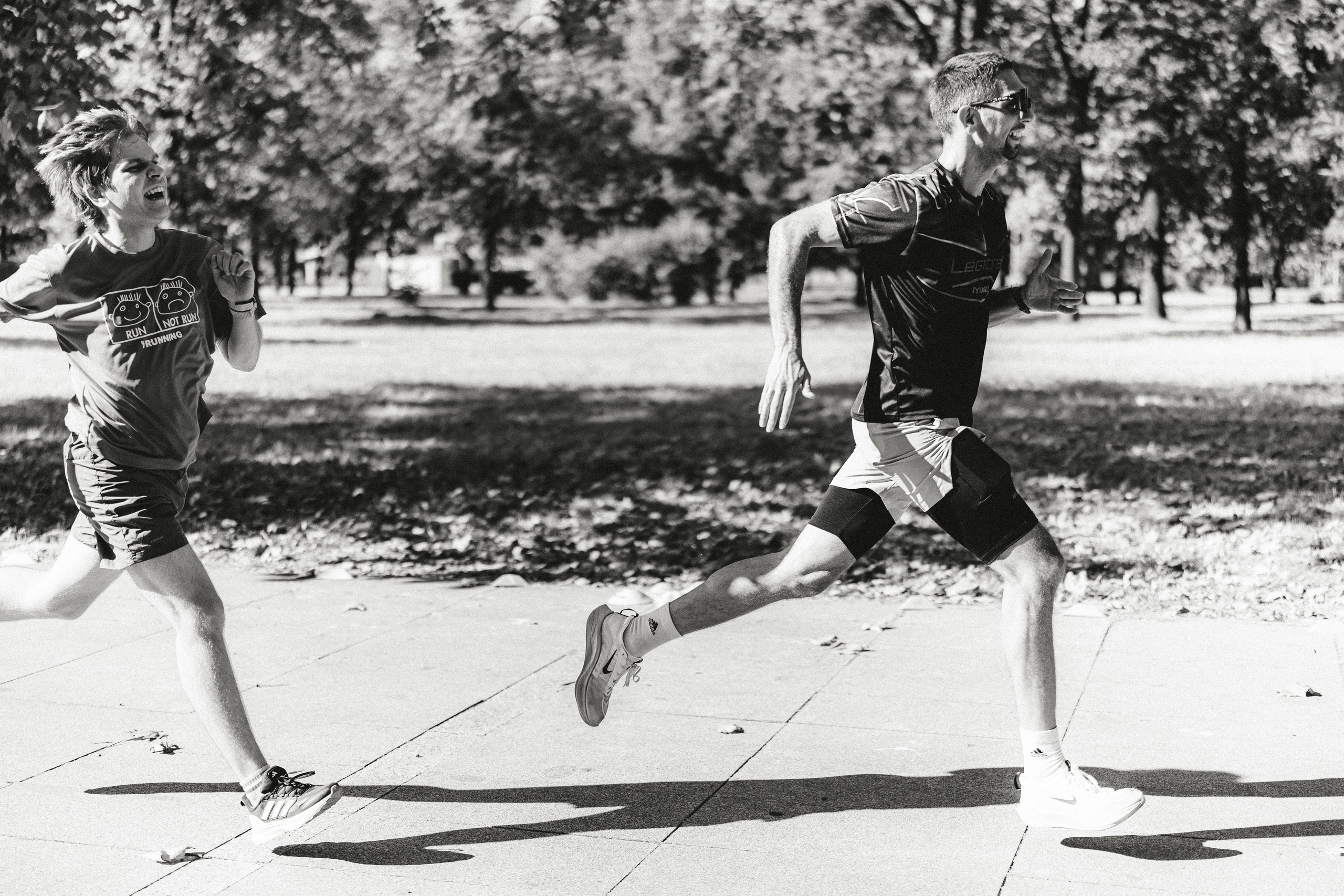 A dynamic black and white action shot capturing two runners mid-stride and laughing during a competitive moment at the S95 park run in Belgrade.